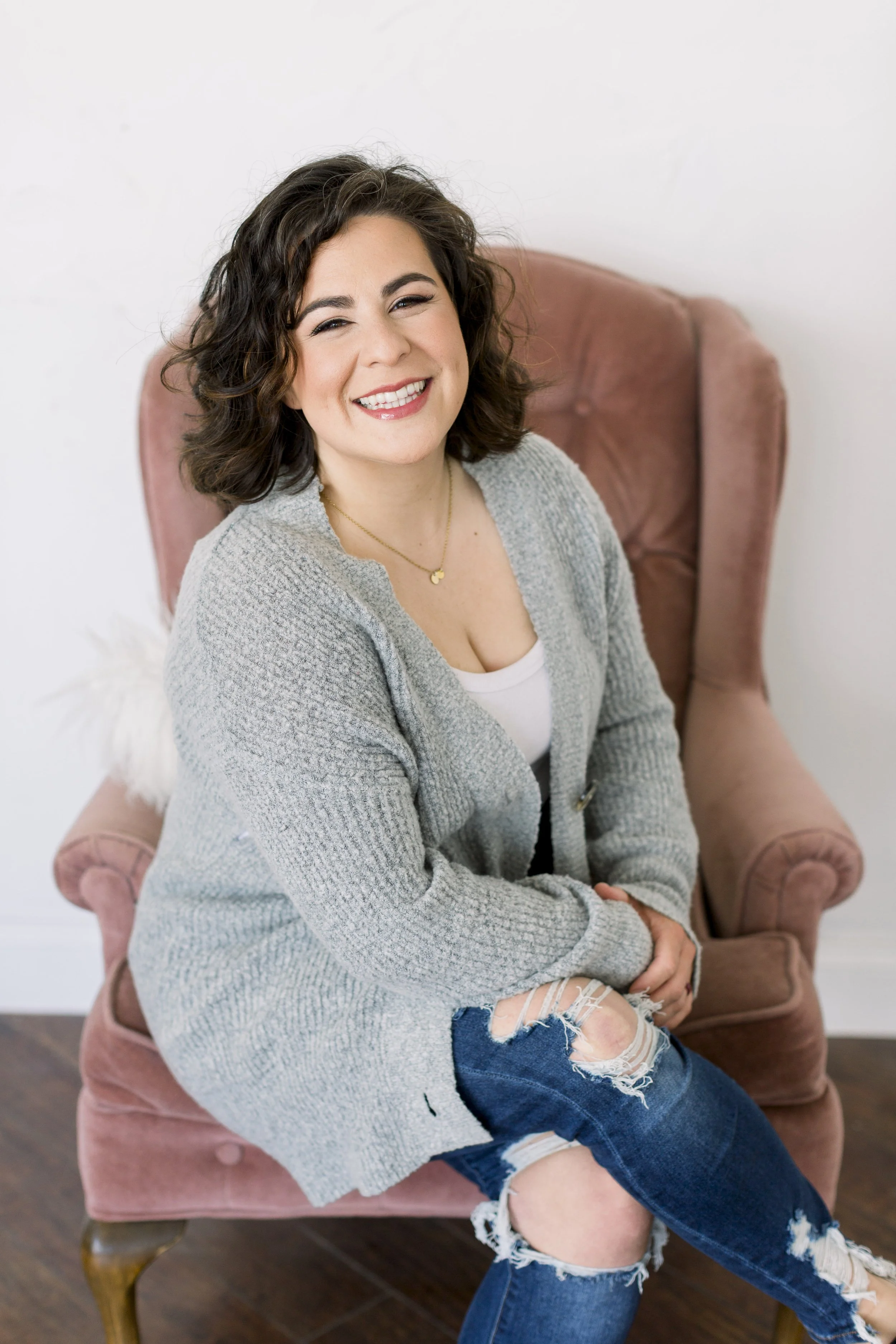 A woman with dark, curly hair smiles while sitting on a pink armchair in a room with white walls and wooden flooring.