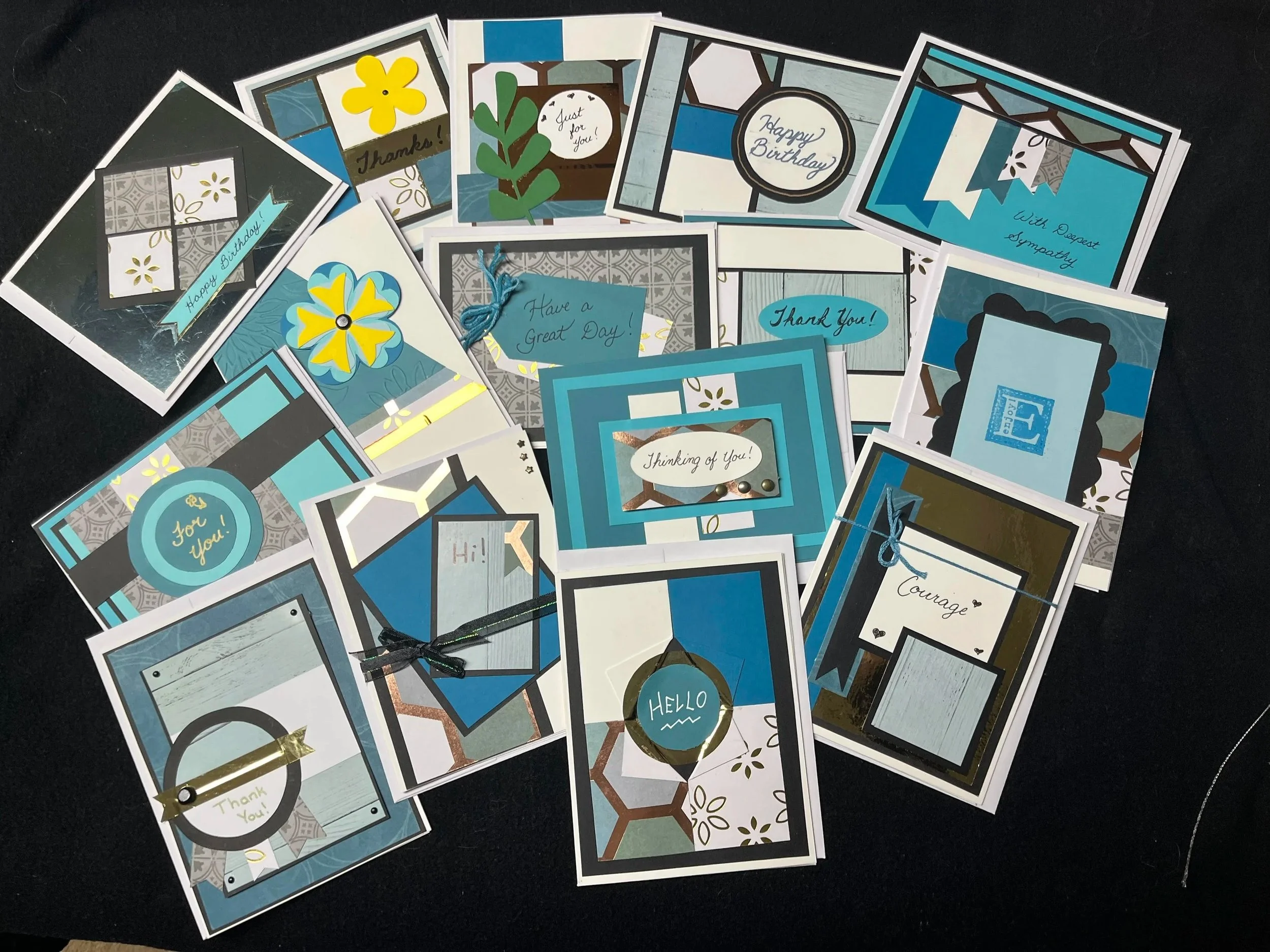 A collection of handmade greeting cards with various designs and messages, mostly in blue, gray, white, and gold, spread out on a black surface.