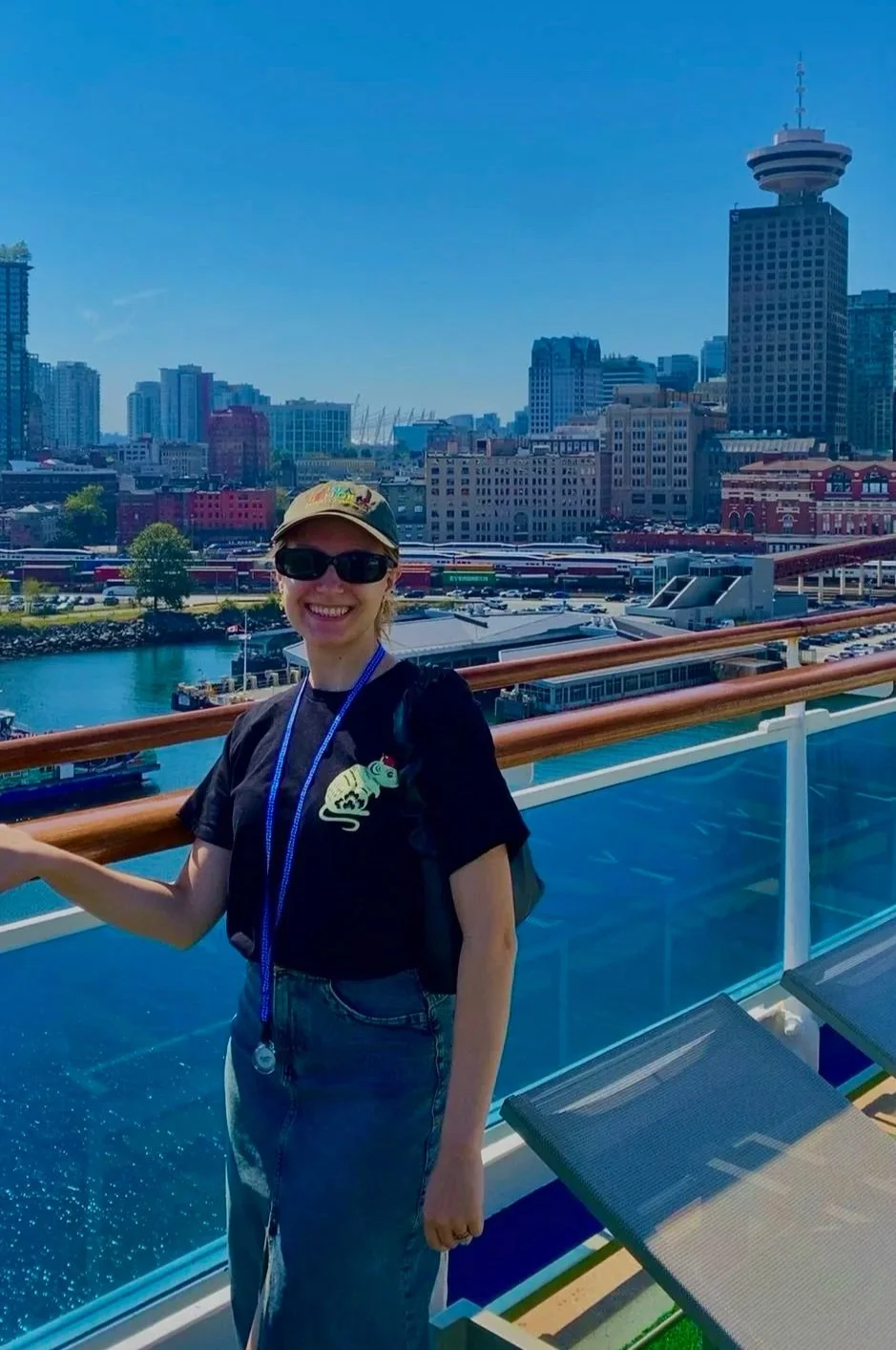 A woman smiling on a cruise ship deck with a city skyline in the background, including tall buildings and a harbor.
