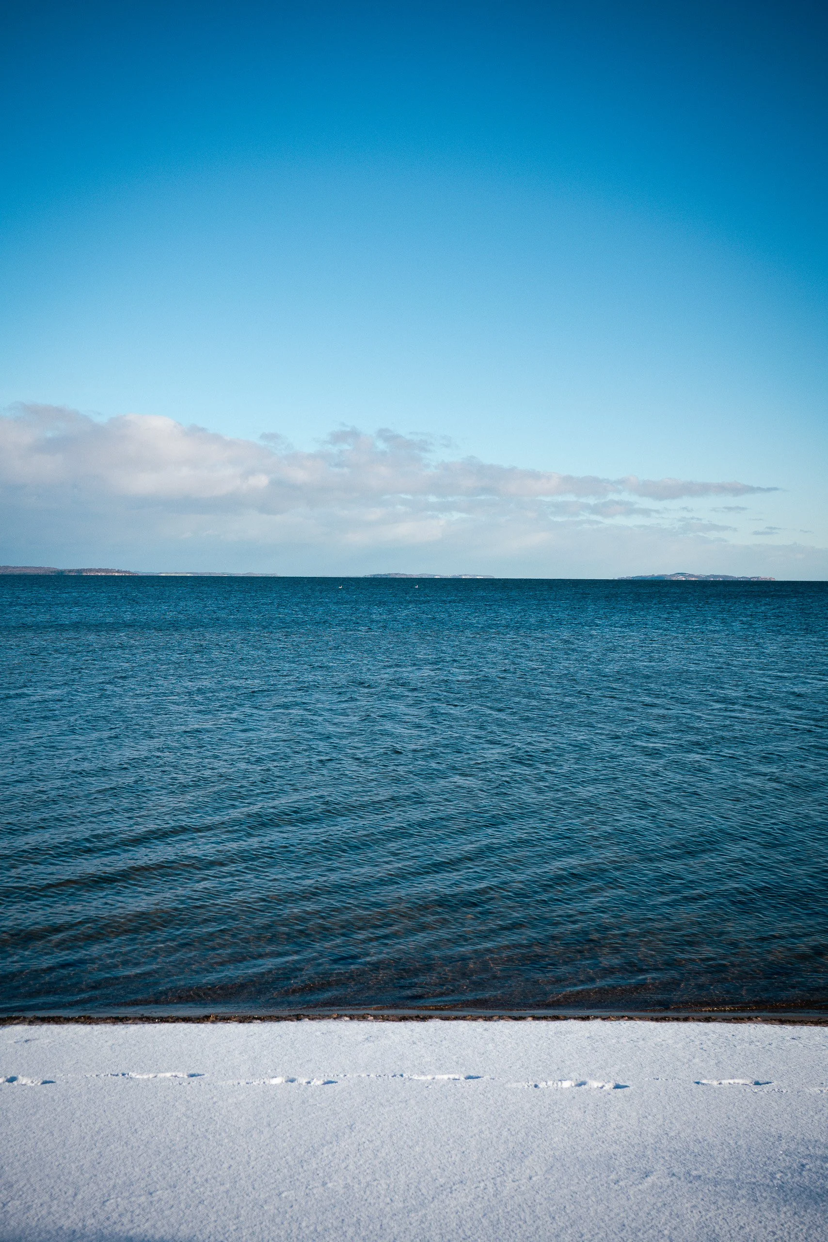 Blick auf das Meer mit einem klaren blauen Himmel, Sandstrand im Vordergrund, leichte Wolken am Himmel.