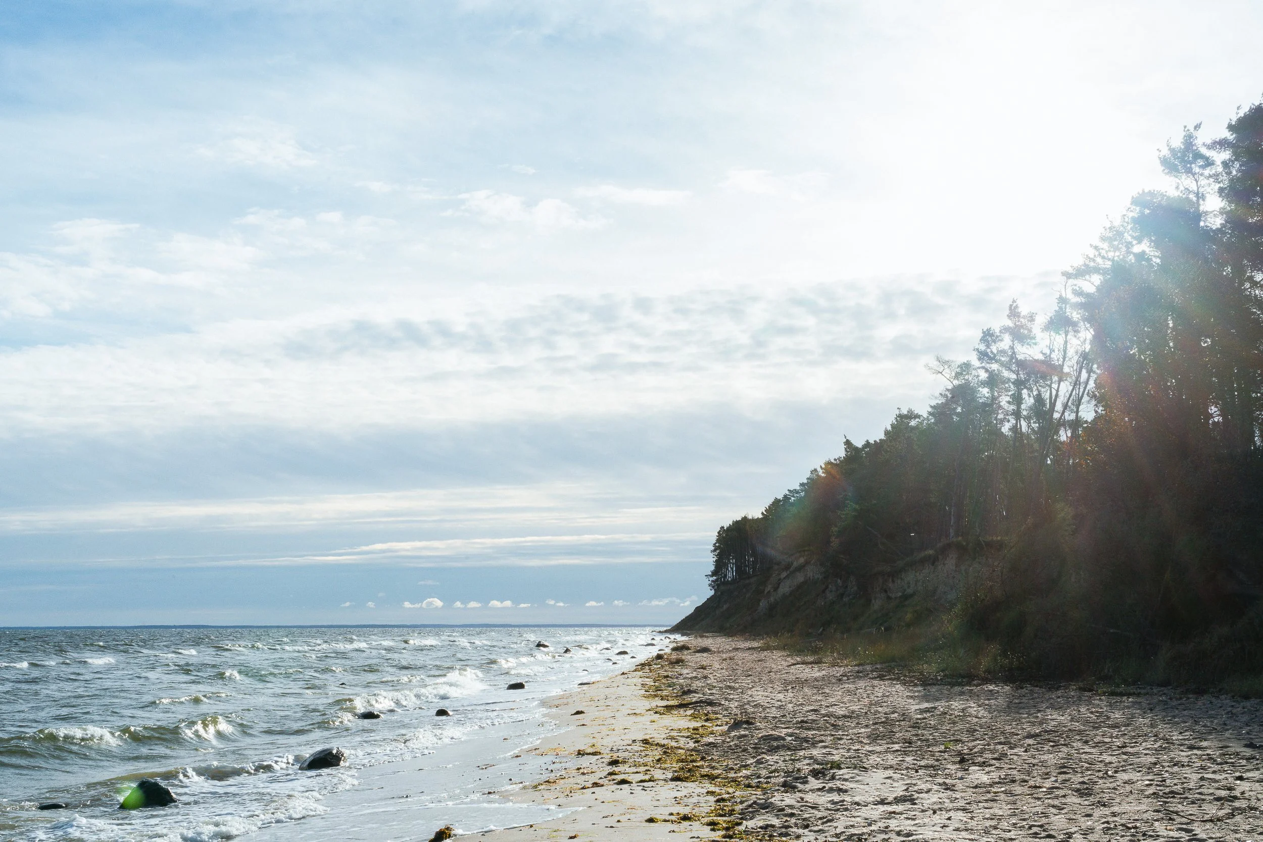Ein Strand mit Sand, Meereswellen und einer Klippe mit Bäumen, Sonnenschein und leicht bewölktem Himmel.