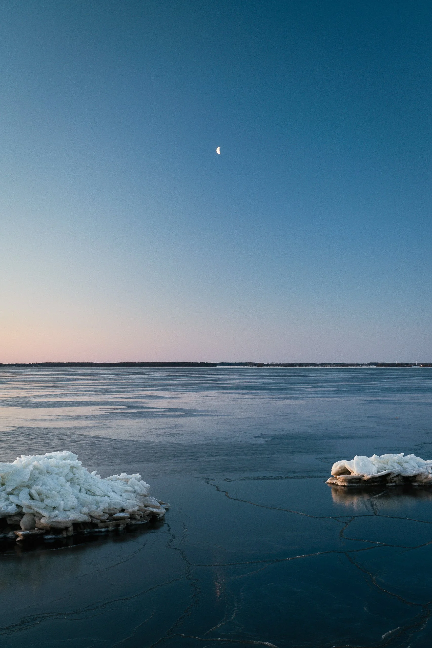 Verschneite Eisschollen auf einem gefrorenen Wasser bei Dämmerung mit Mond am Himmel.