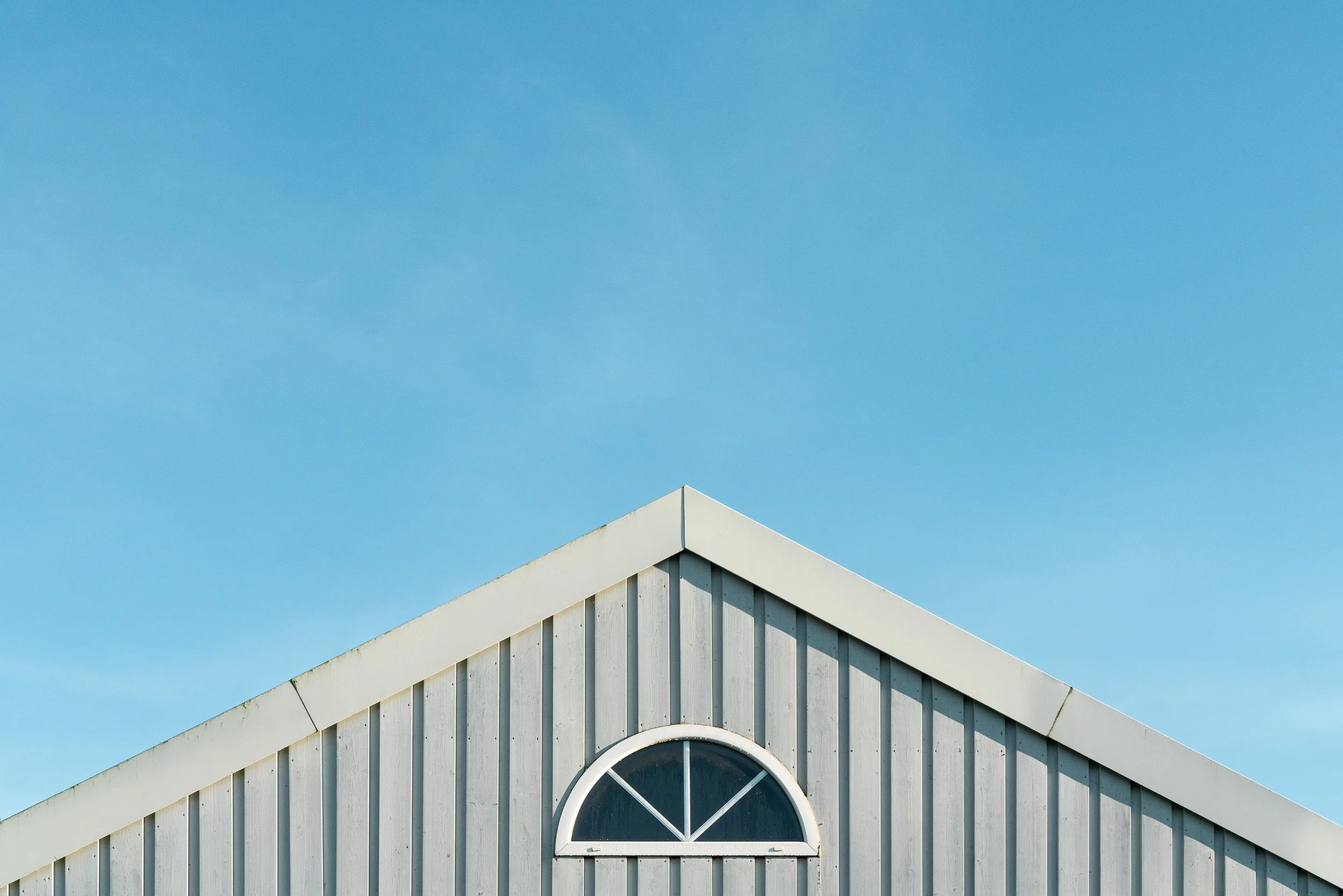 Das Dach eines Hauses mit weißen Holzpaneelen und einem halbkreisförmigen Fenster unter einem blauen Himmel.