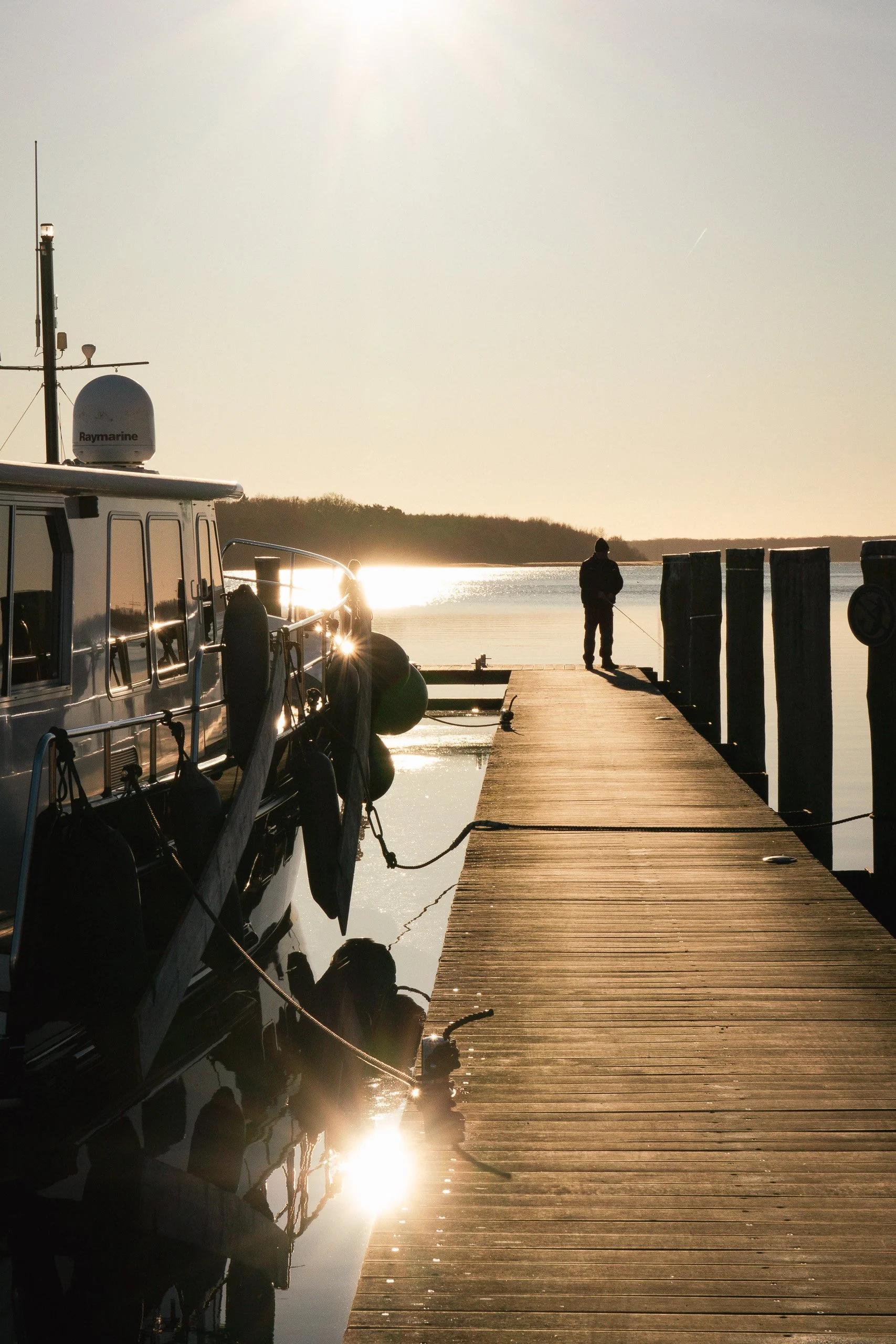 Ein Boot ist an einen Steg am Wasser befestigt, während eine Person im Hintergrund angelt und die Sonne untergeht.