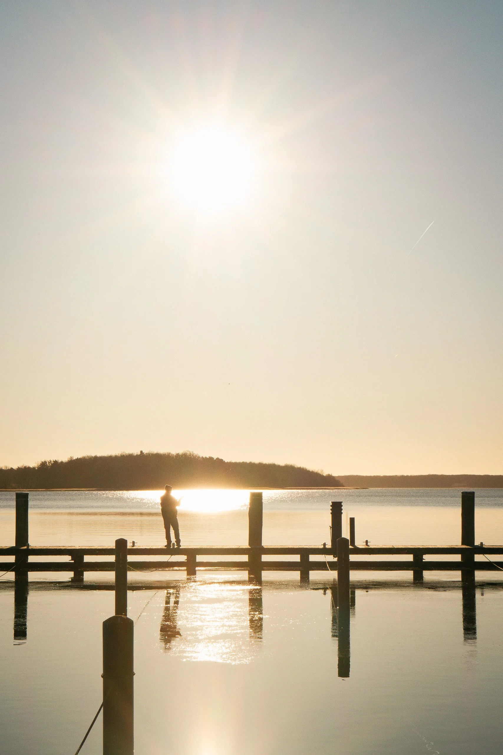 Silhouette eines Mannes, der auf einem Steg bei Sonnenuntergang am Wasser steht, mit reflektierendem Wasser und einem bewölkten Himmel im Hintergrund.