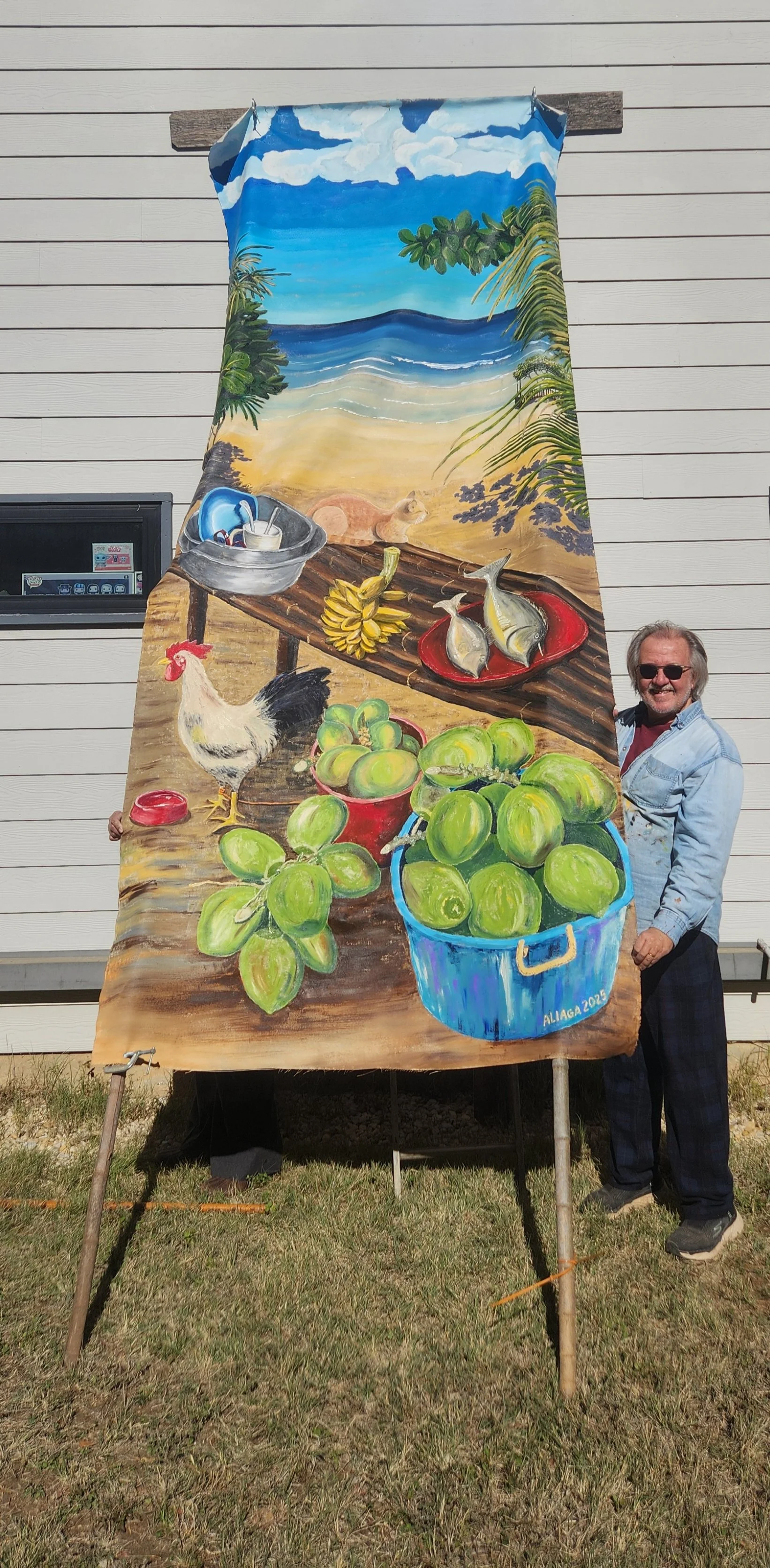 A man wearing sunglasses and a light blue shirt stands beside a large painted outdoor banner depicting a beach scene with fresh produce and seafood, including green apples in a blue bucket, bananas, fish on a platter, and chickens, with a sandy beach, ocean, and sky with clouds in the background.