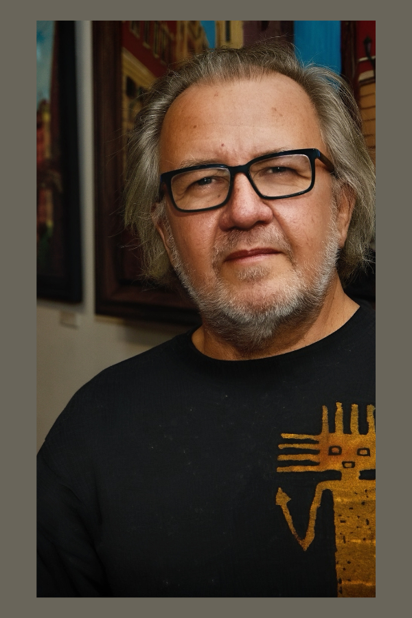 A middle-aged man with gray hair and beard wearing black glasses and a black T-shirt with a yellow design, standing in front of a colorful background with artwork.