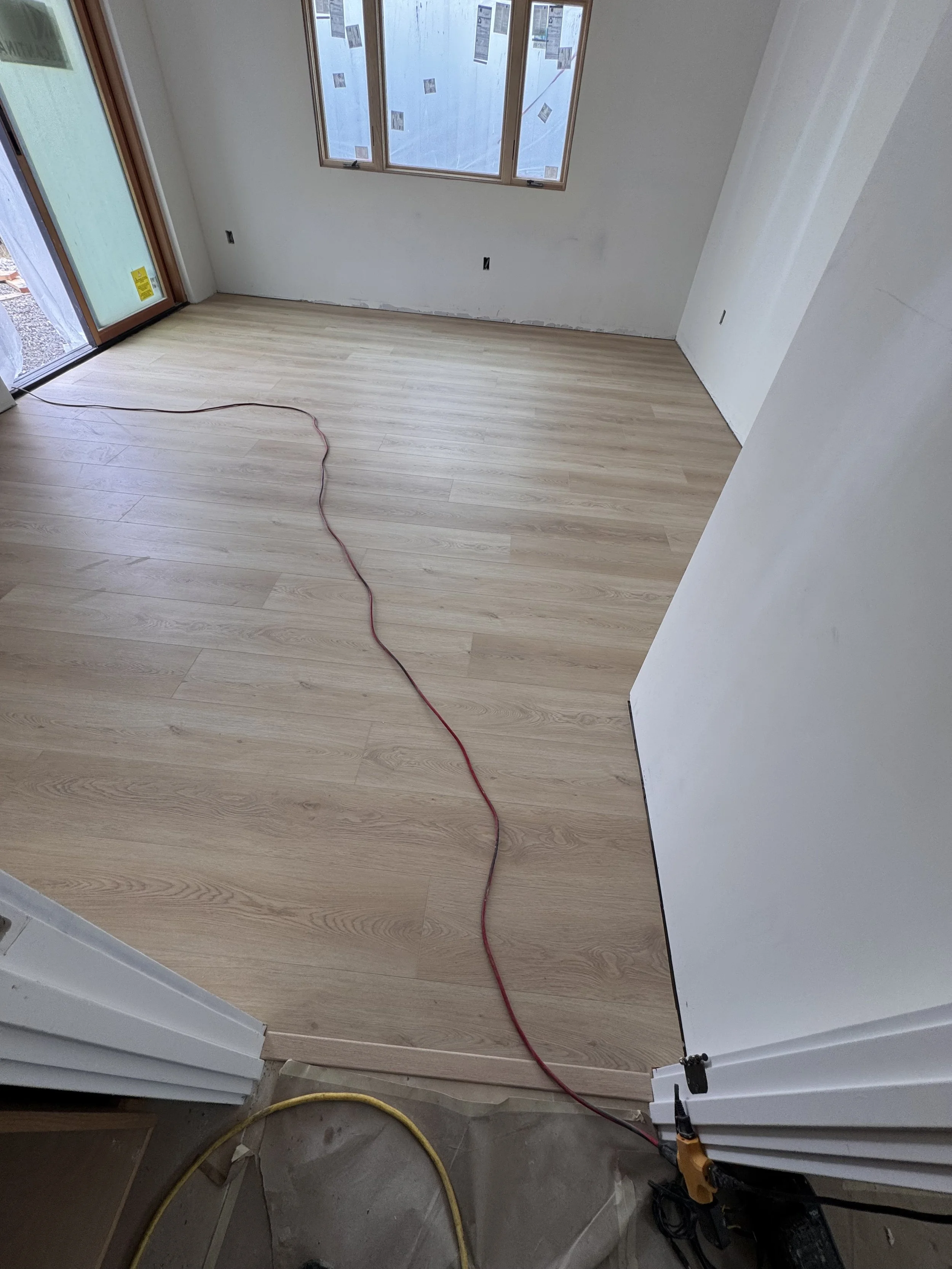 An unfinished room with newly installed light-colored wooden flooring, white walls, and large windows covered with protective plastic. Construction tools and electrical cords are visible, indicating ongoing renovation.