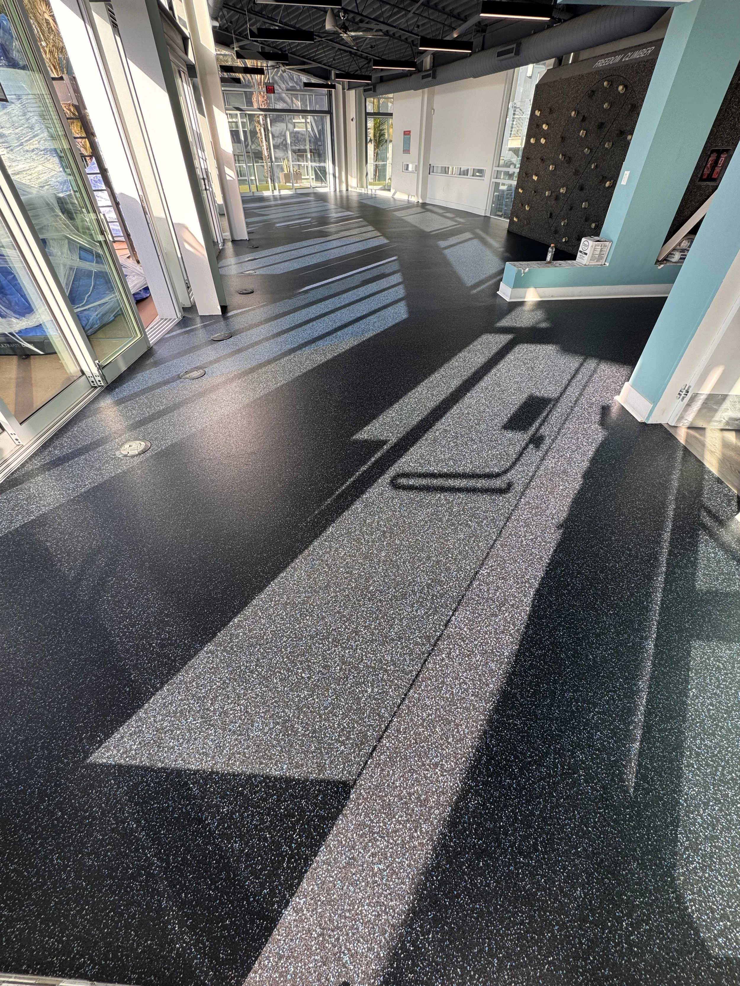 Empty modern lobby with shiny black and speckled gray flooring, sunlight streaming through large glass doors, and a climbing wall on the right wall.