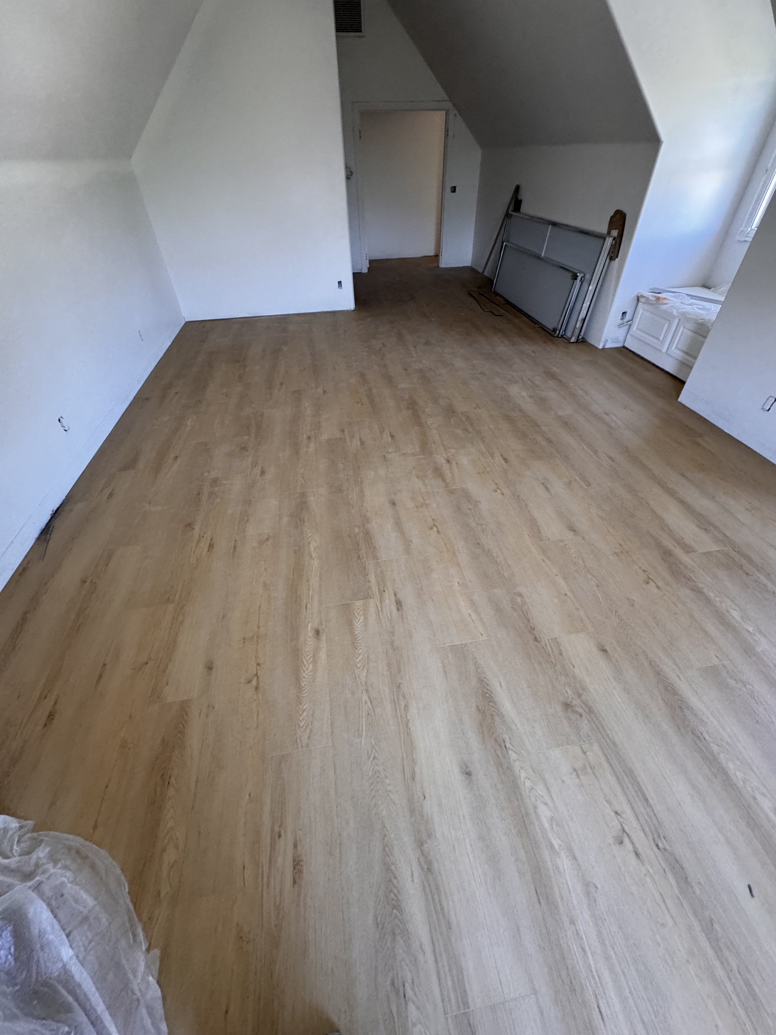 Empty room with newly installed light wood flooring, white walls, a window, and some unfinished construction materials leaning against the wall.