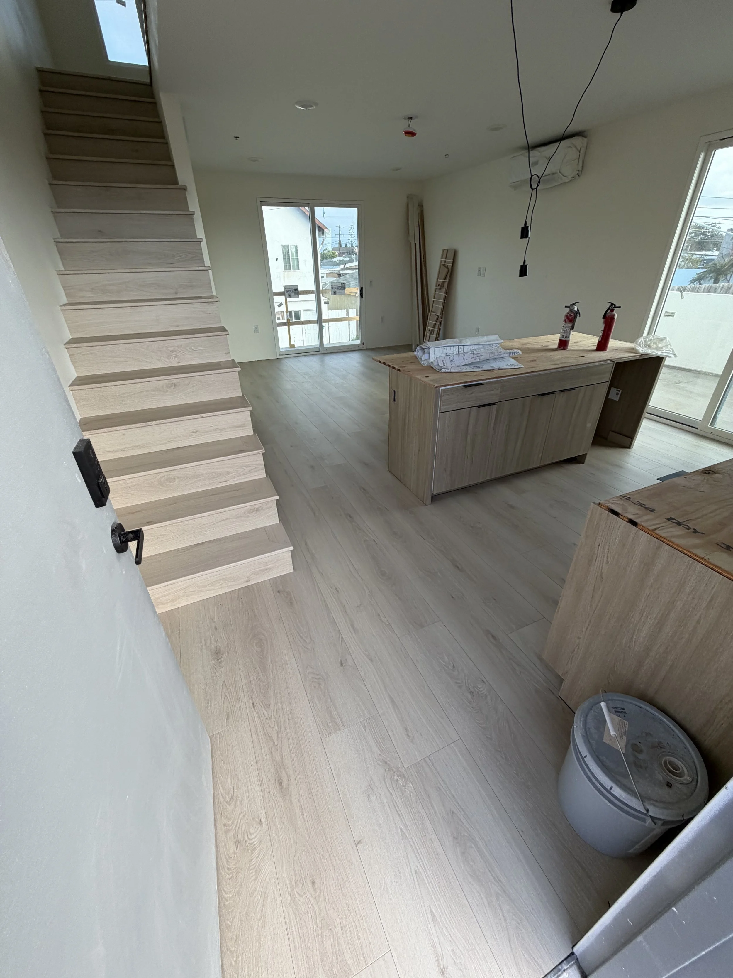 Interior of a house under construction with light wood flooring, wooden stairs, a kitchen island, and sliding glass doors leading outside.