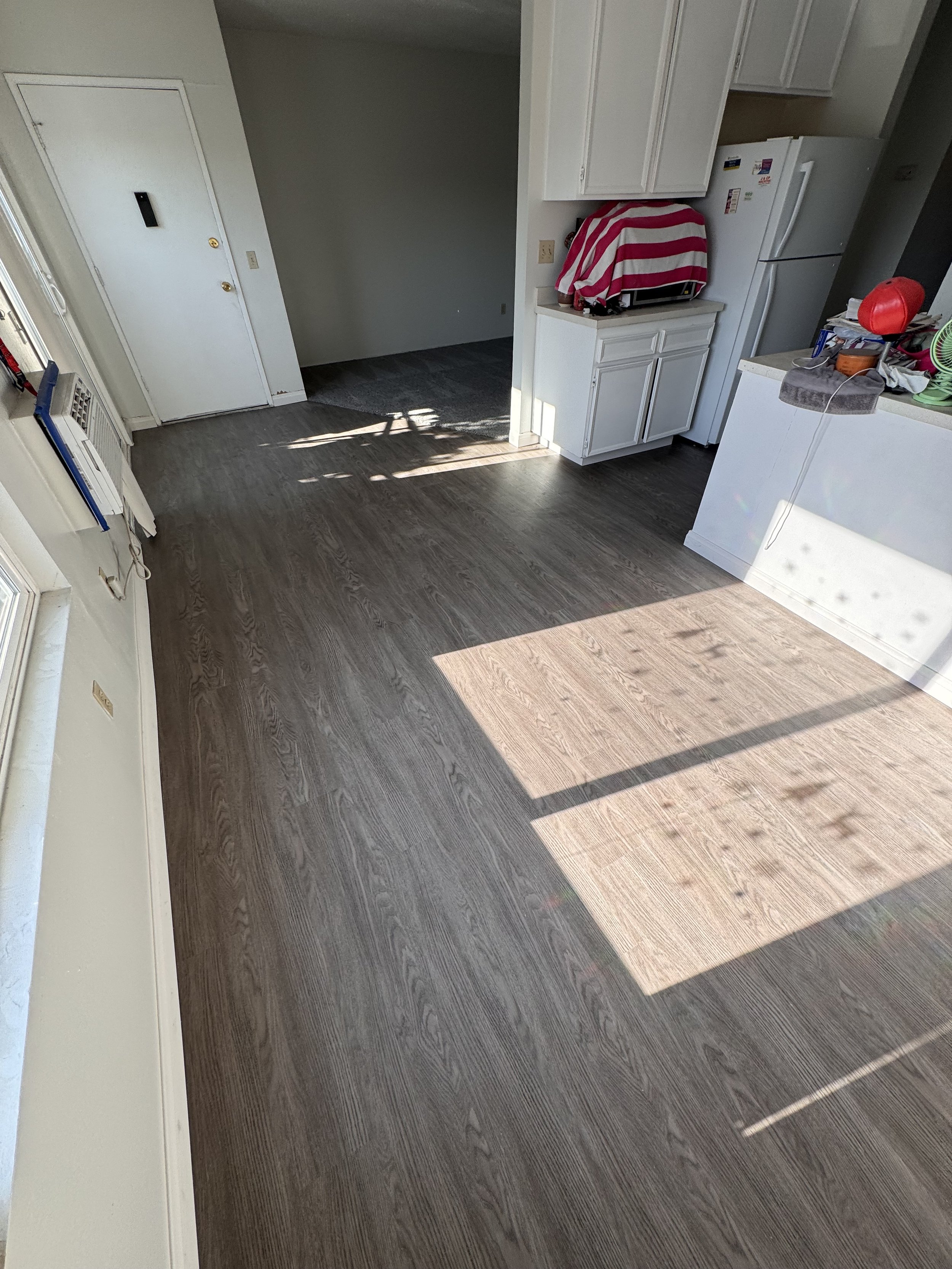 Empty kitchen with dark wood flooring, white cabinets, and a white refrigerator covered with a red and pink striped cloth. Sunlight streams through a window, creating a pattern on the floor.
