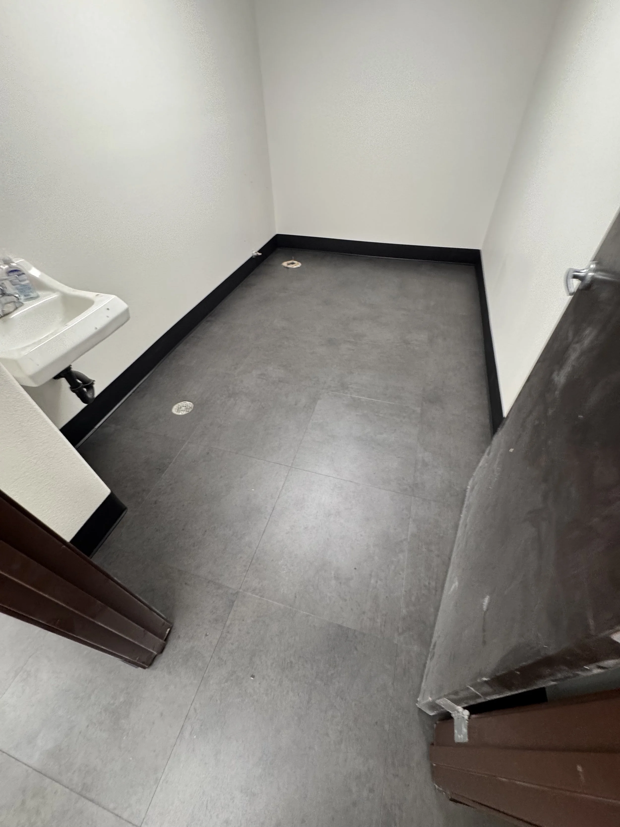 Empty small room with white walls, a small sink in the corner, and black baseboards. The floor has gray tiles, and the room appears to have plumbing fixtures for a bathroom or utility space.