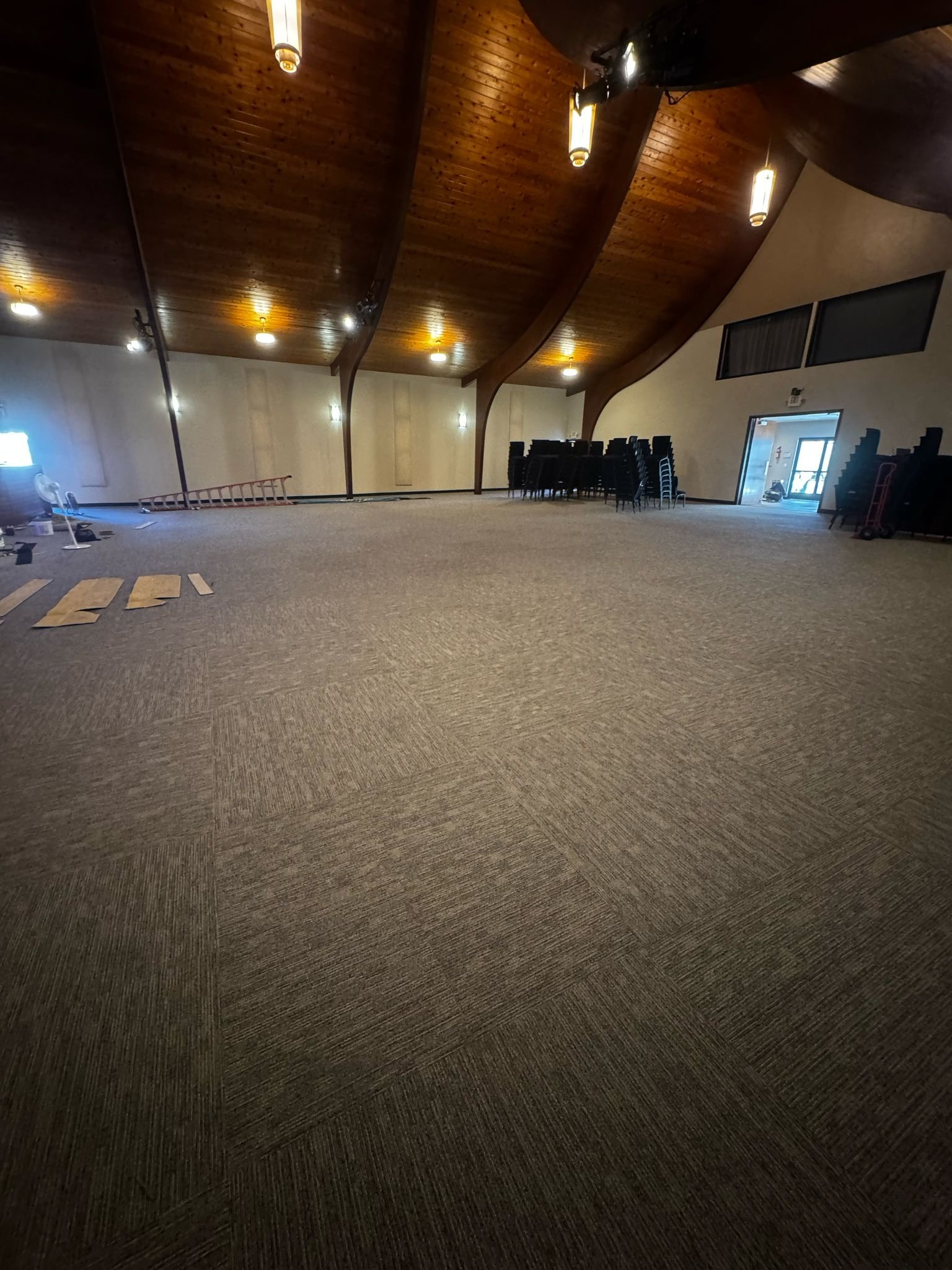 Empty room with a high wooden ceiling, beige walls, and carpeted floor, with stacked chairs and some equipment along the walls.