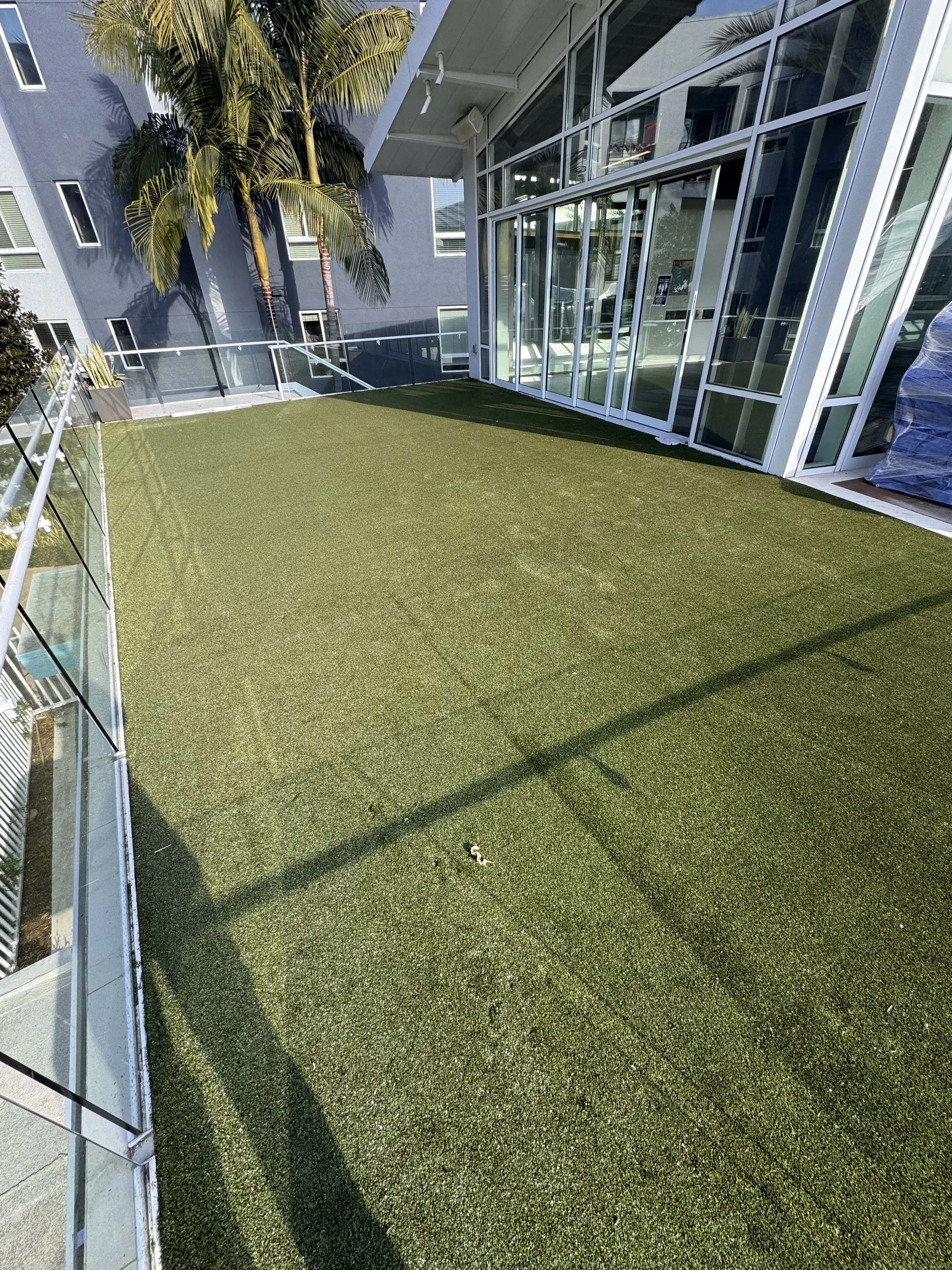 A balcony with artificial grass flooring, glass railing, and a large glass wall with multiple sliding doors, overlooking a neighboring building and palm trees.