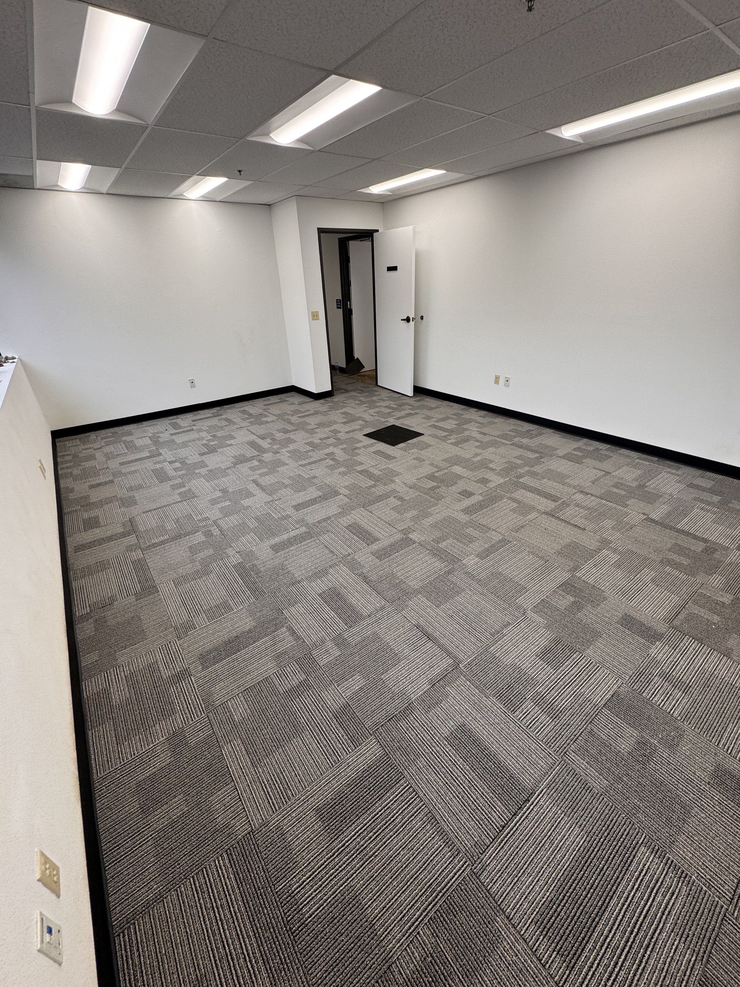Empty office room with patterned carpet, white walls, ceiling with fluorescent lighting, open door leading to hallway.