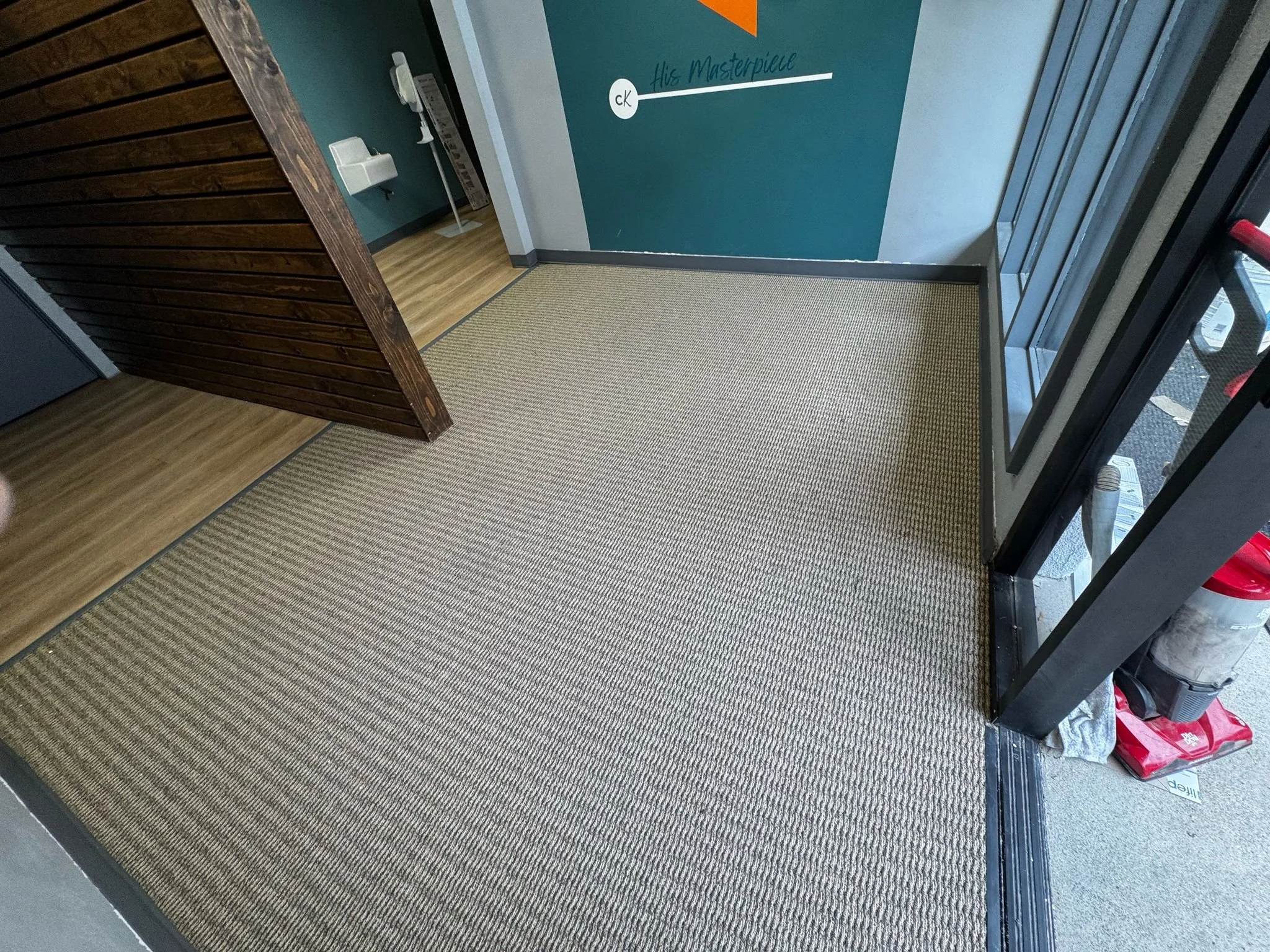 Interior of an entrance with a wedding-themed message on the wall, gray walls, a glass door, and a tight-mesh brown rug.