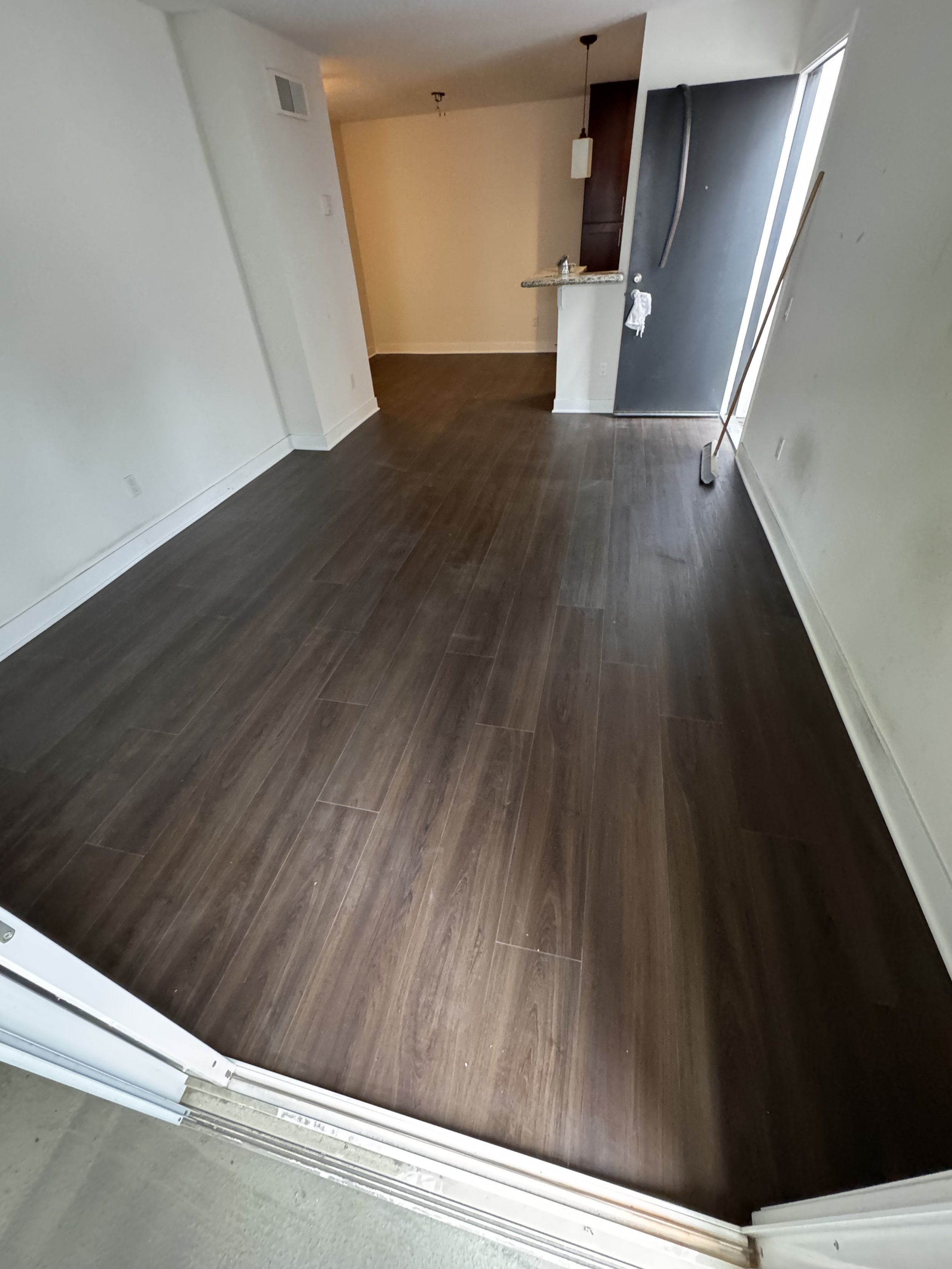 Empty living room with dark wood flooring, white walls, and a small kitchen area in the background with a granite countertop and hanging pendant light.