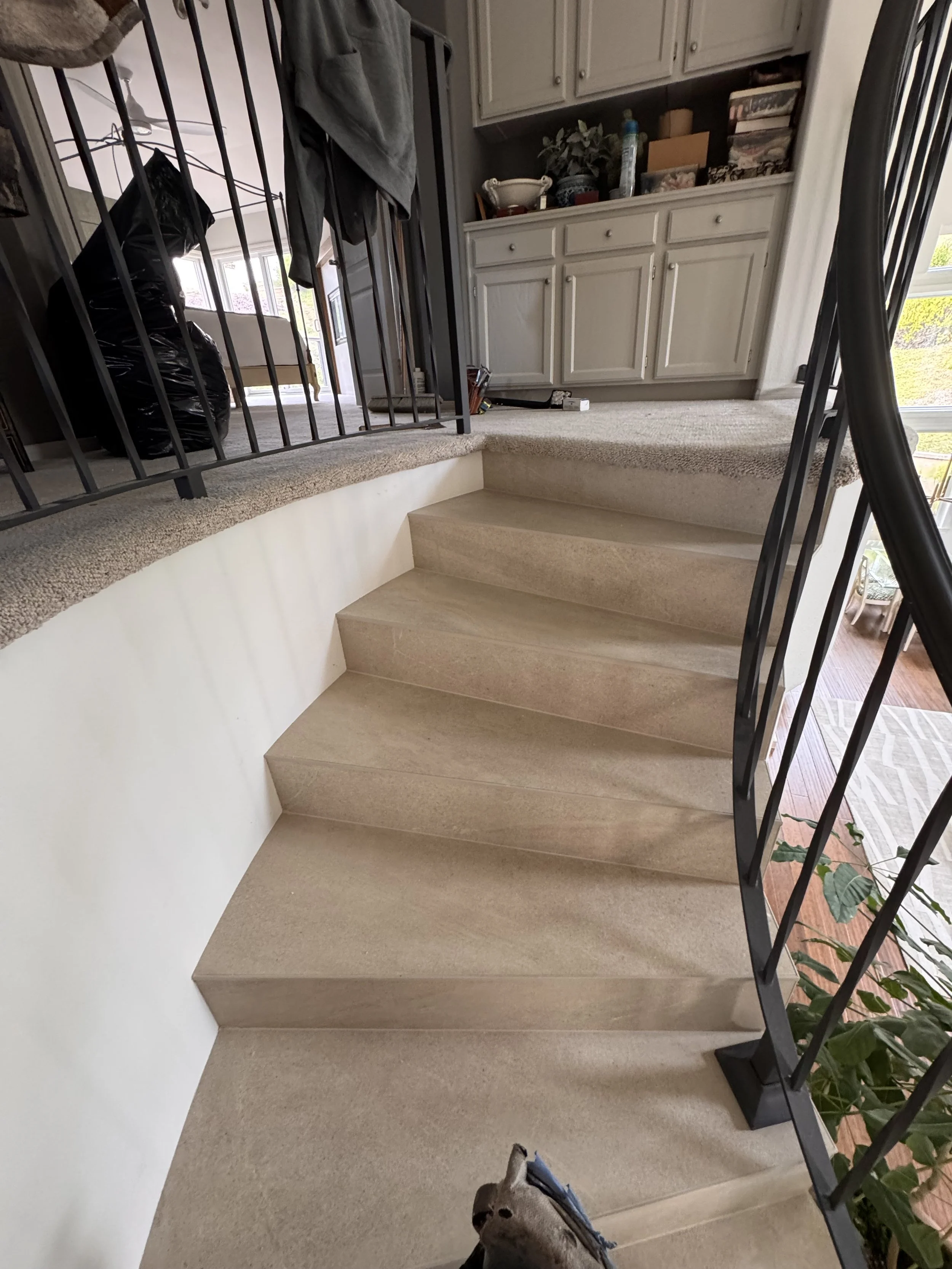 Interior view of a staircase with beige steps and black railing, leading up to a carpeted landing in a home. In the background, there are kitchen cabinets, a countertop with various items, and a window allowing natural light to enter.