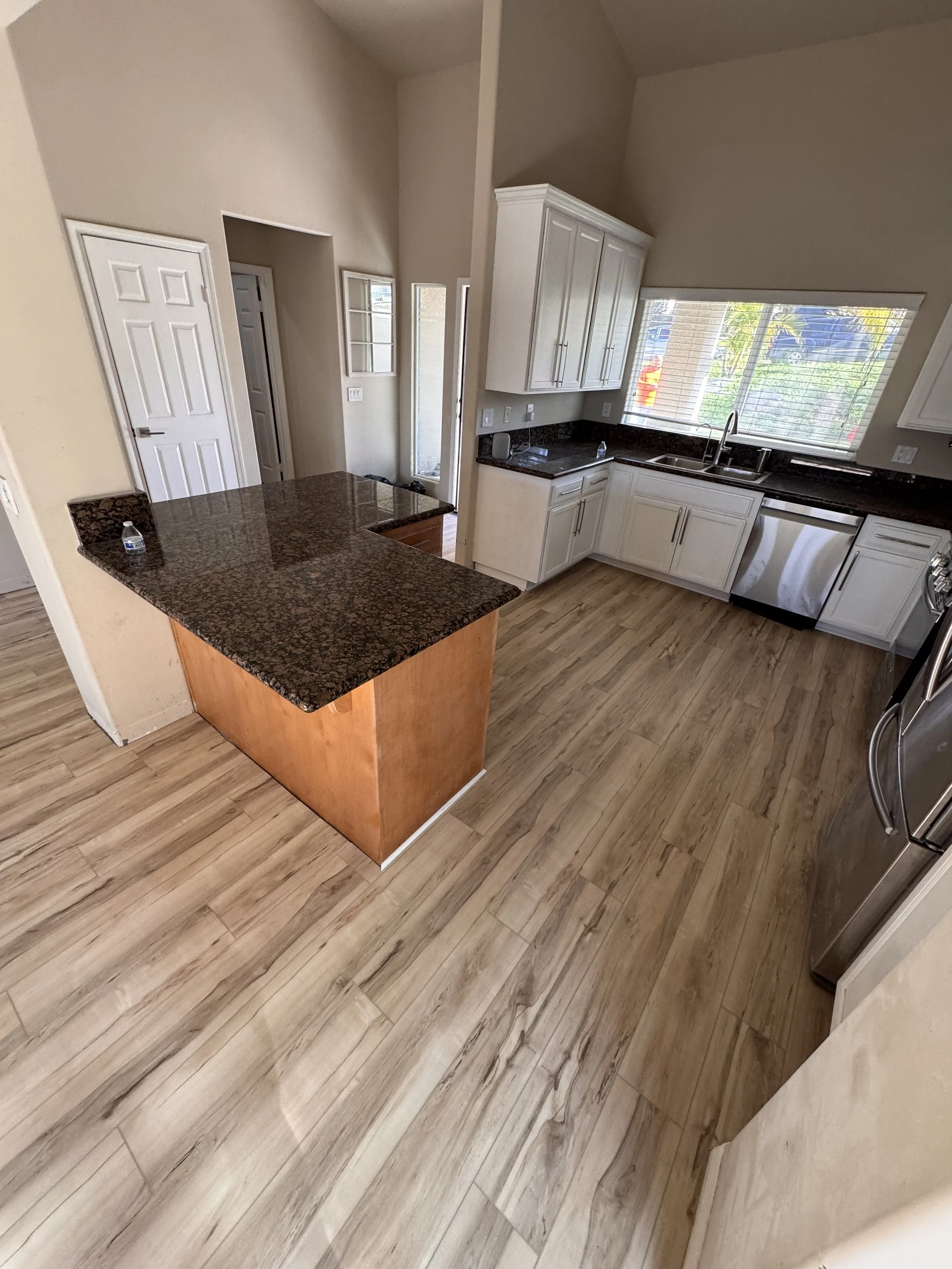 Kitchen with white cabinets, black granite countertops, a stainless steel dishwasher, and a large window above the sink letting in natural light. There is a small island with a dark granite top and a wooden base.