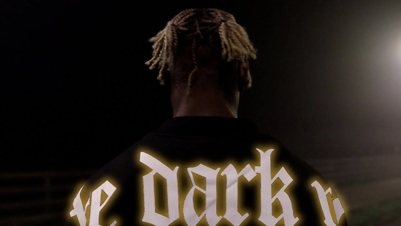 Back view of a person with blonde dreadlocks wearing a black jacket with glowing white text that reads "the dark".