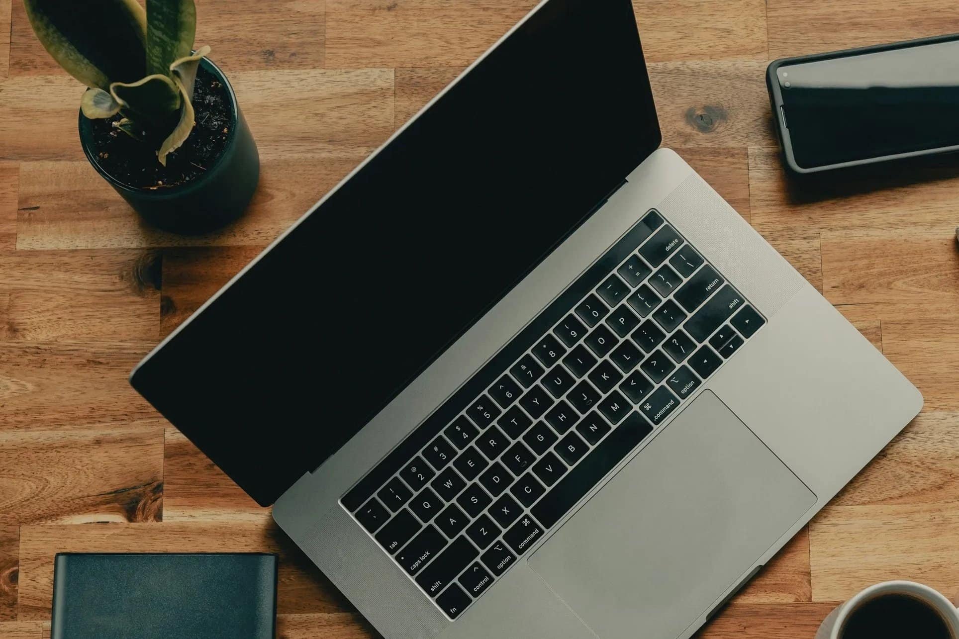 Top-down view of a wooden desk with a silver laptop, a black smartphone, a green and yellow potted plant, a black notebook, and a white mug.