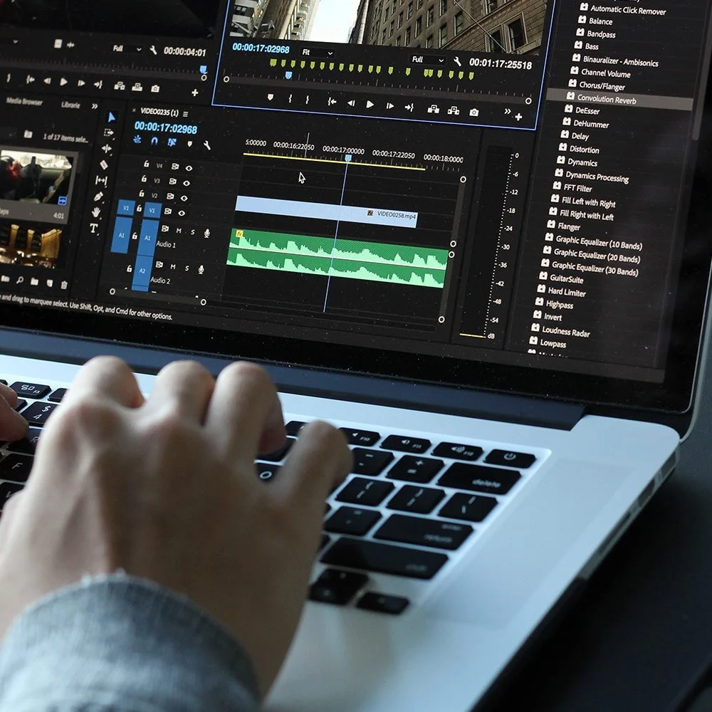 Close-up of a person's hand using a laptop with a photo and video editing software on the screen, showing audio tracks, effects, and a cityscape reflection on the monitor.