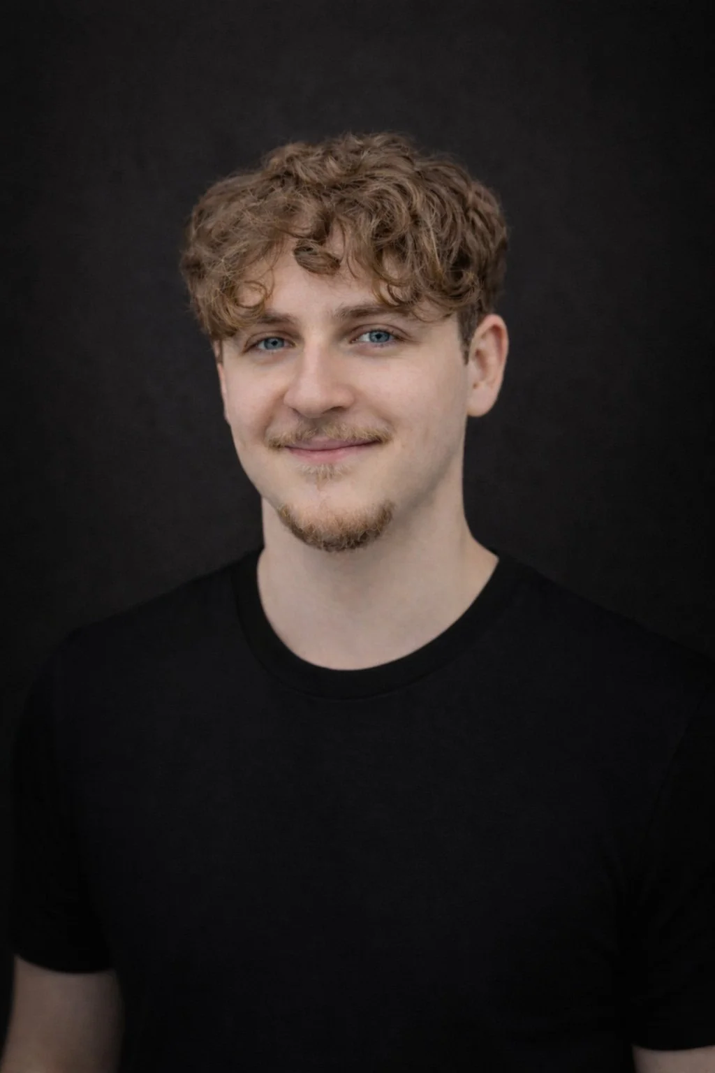 Portrait of a young man with curly light brown hair, blue eyes, and a light beard, wearing a black shirt, smiling, against a dark background.