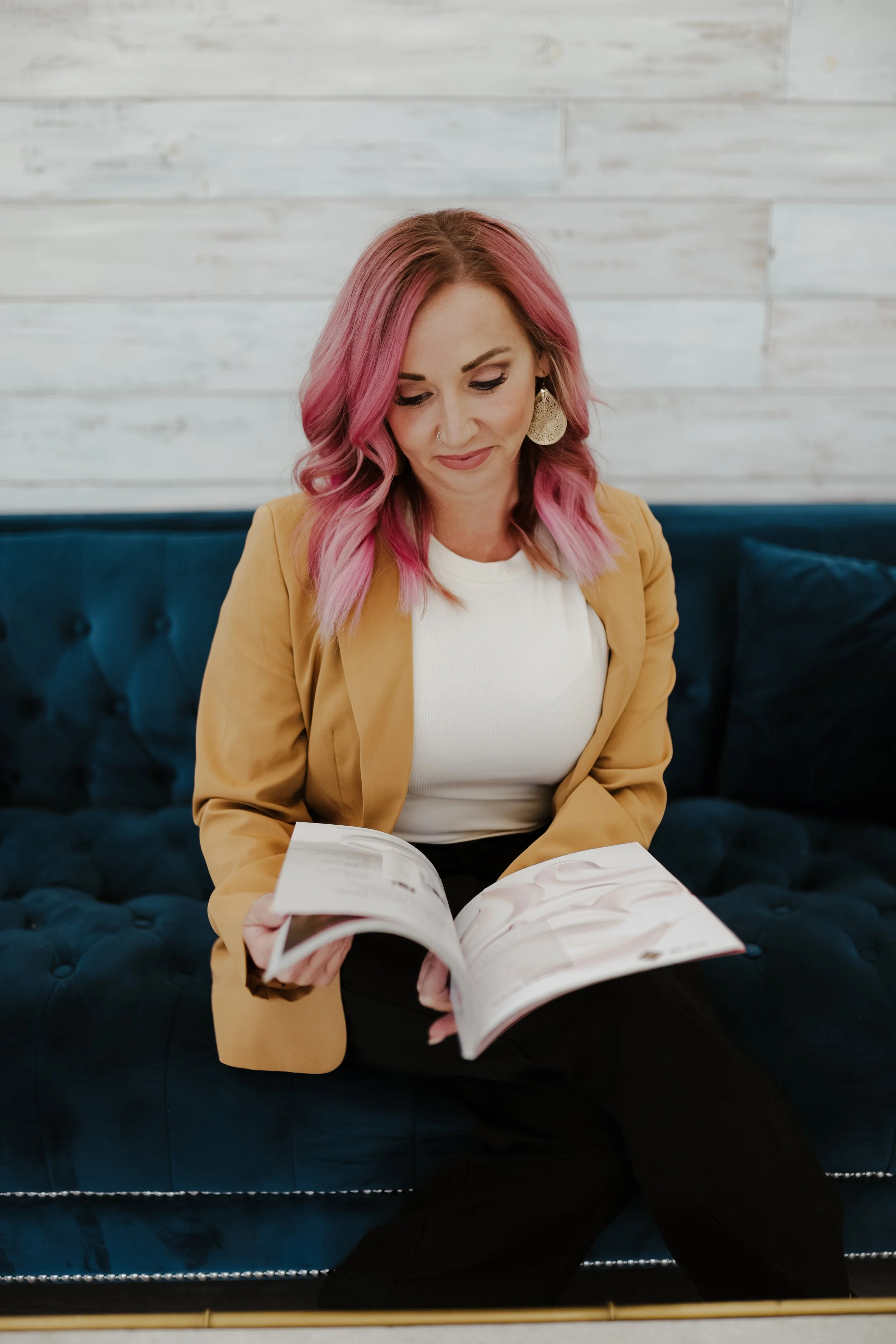 A woman with pink hair, wearing a tan blazer and white shirt, sitting on a dark blue velvet couch, looking at an open magazine.