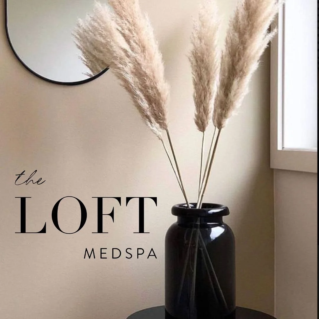 A black vase with three large pampas grass stems on a small black table next to a round mirror on a beige wall and a window.