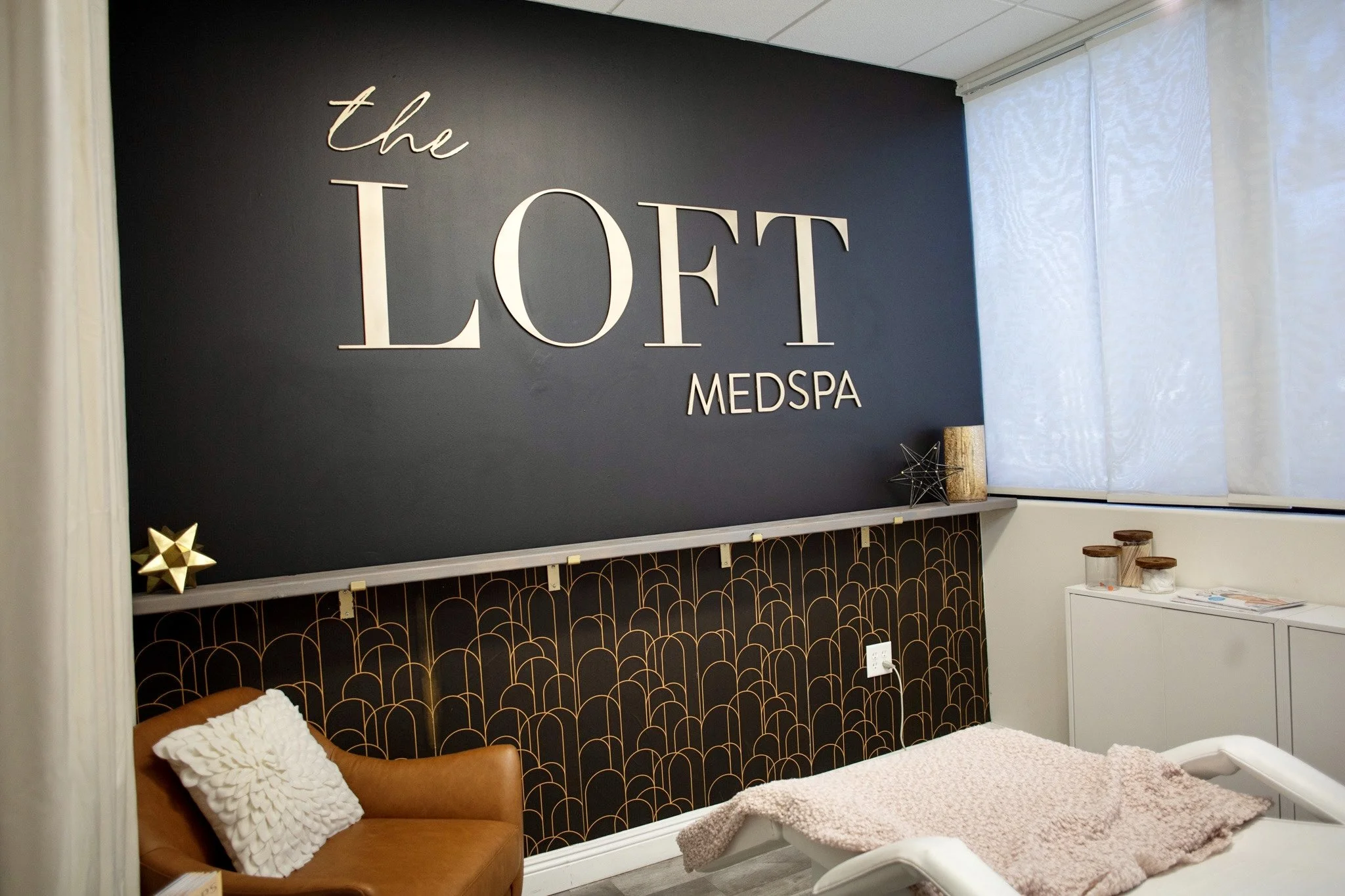 Interior of a medspa with a large sign on the wall that says 'the LOFT MEDSPA'; includes a brown leather chair with a white pillow, a bed with a pink blanket, and decorative objects on a shelf near a window with white blinds.