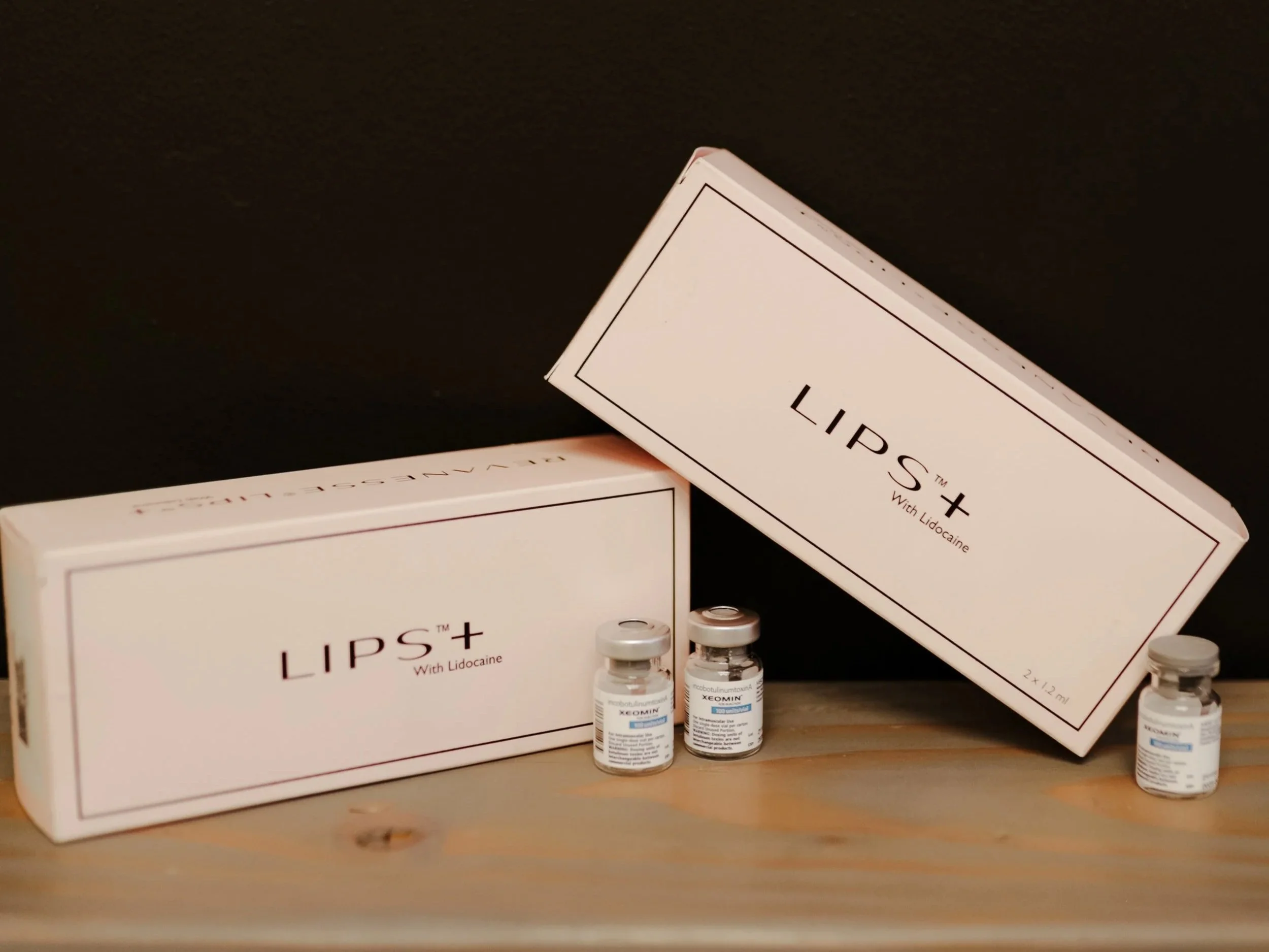 Two boxes labeled 'LIPS+' with lidocaine and two small vials of medication are placed on a wooden surface against a dark background.