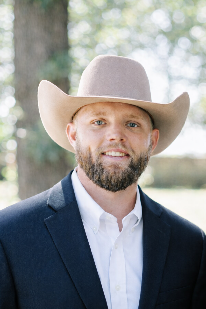A man wearing a large cowboy hat, dark blazer, and white shirt, smiling outdoors with blurred trees in the background.