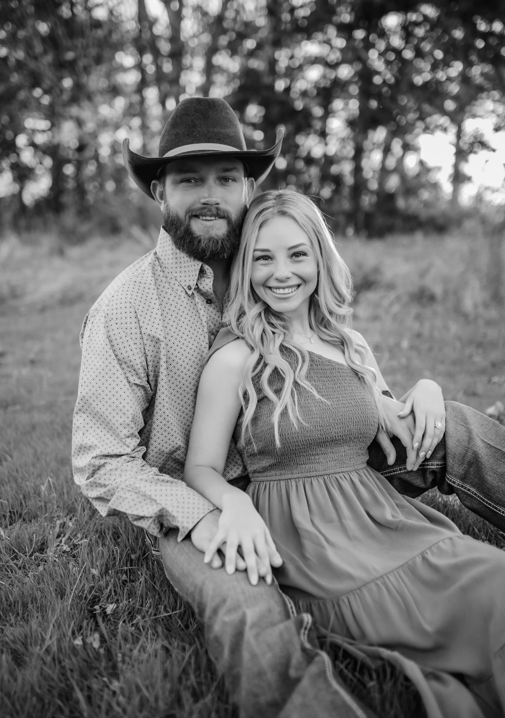 A smiling couple sitting on the grass outdoors, with the man wearing a cowboy hat and the woman in a sleeveless dress, both looking at the camera.