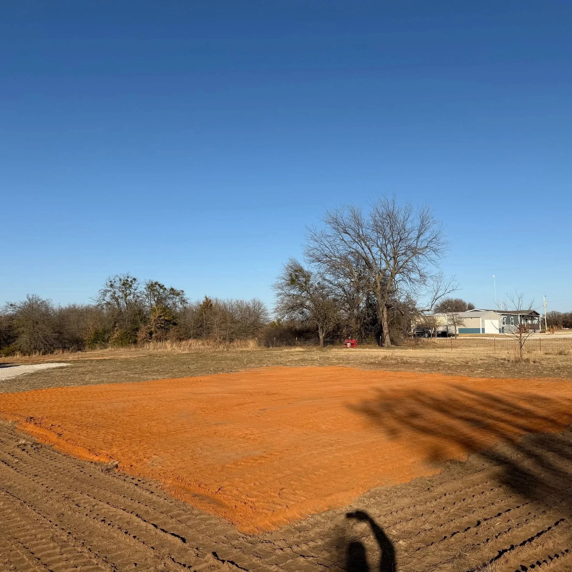 Shows a newly built building pad, surrounded by leafless trees and a house in the background under a clear blue sky.