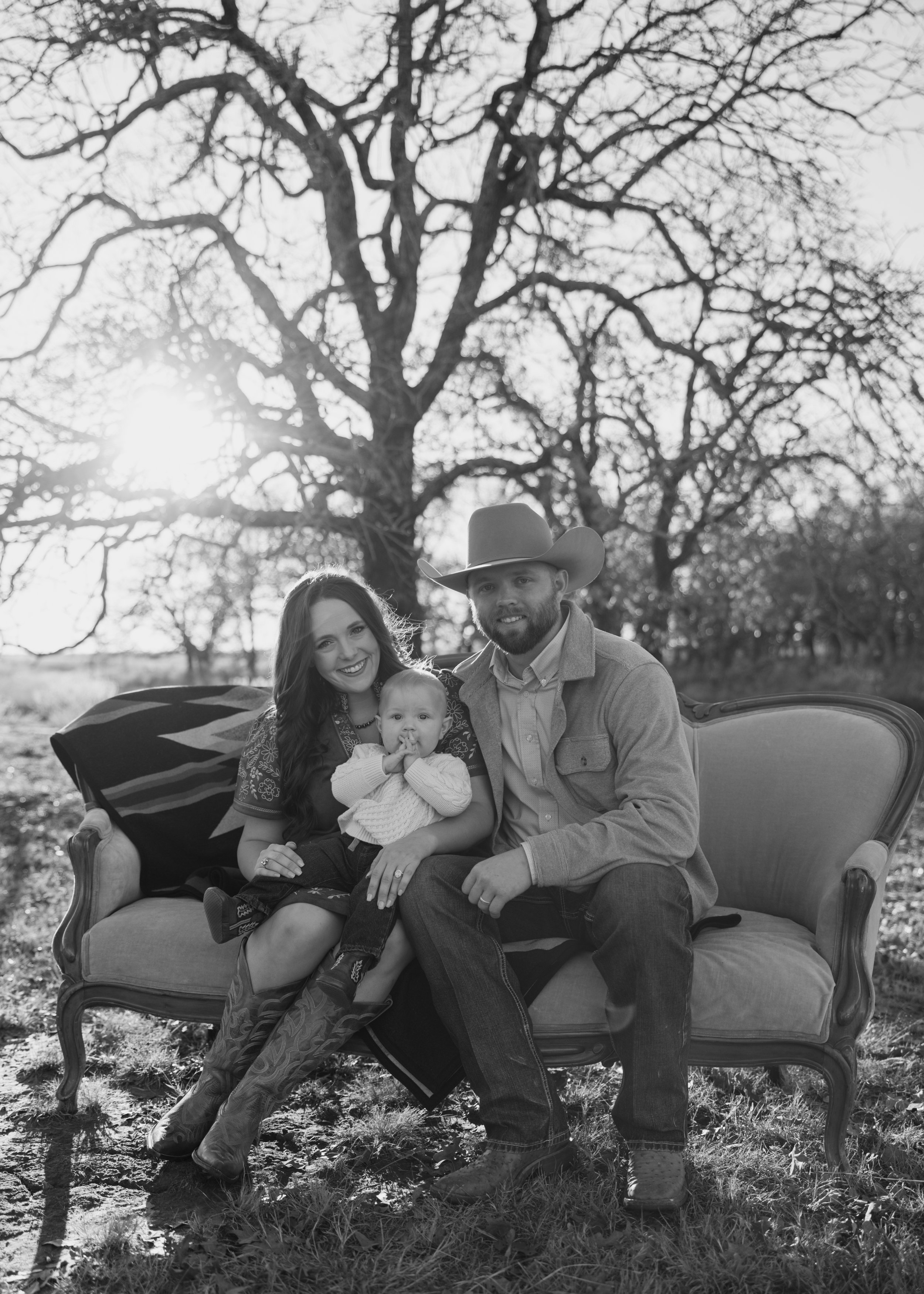 A black and white photo of a family sitting on an outdoor vintage sofa in a park, with a large tree and sun in the background. The family includes a woman, a man wearing a cowboy hat, and a young child.