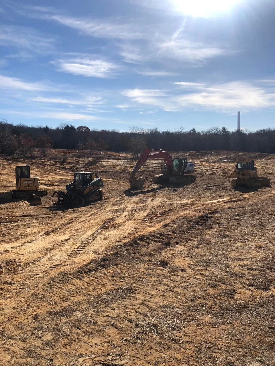 Four construction vehicles, including excavators and bulldozers, parked on a dirt construction site or a pond construction site under a partly cloudy sky.
