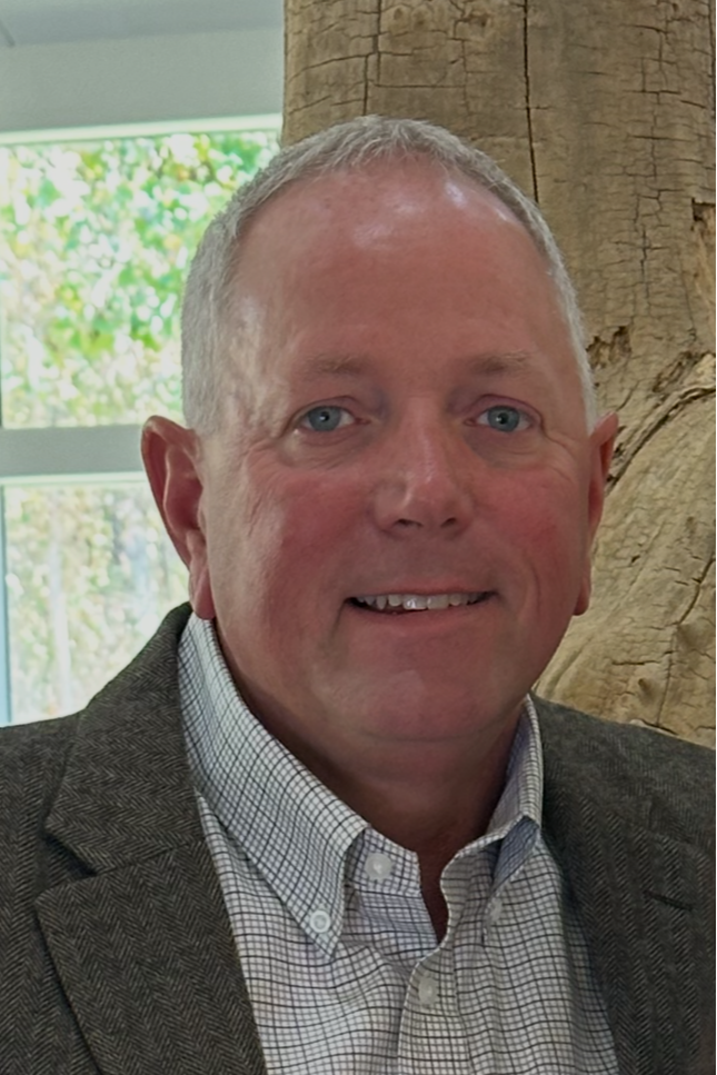 A middle-aged man with short gray hair, blue eyes, and a slight smile, wearing a gray blazer and a checked dress shirt, standing indoors in front of a tree trunk and window with greenery outside.