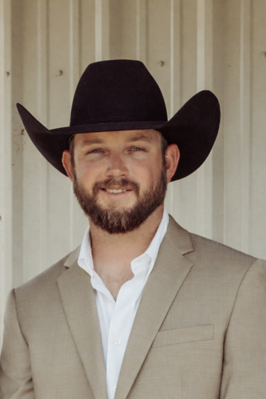 A man wearing a black cowboy hat, light beige blazer, and white shirt, standing outdoors against a light-colored metal wall.