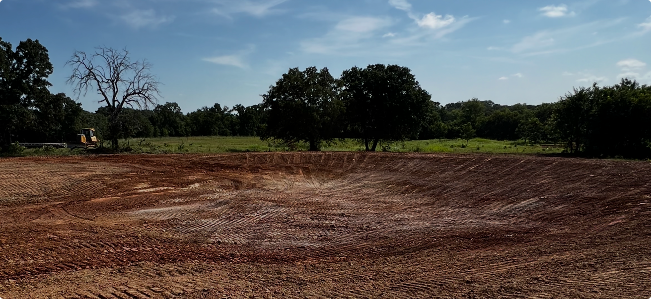 A large dirt excavation site or pond constructionsite with a dozer and trackhoe in the distance, surrounded by trees and open fields under a partly cloudy sky.