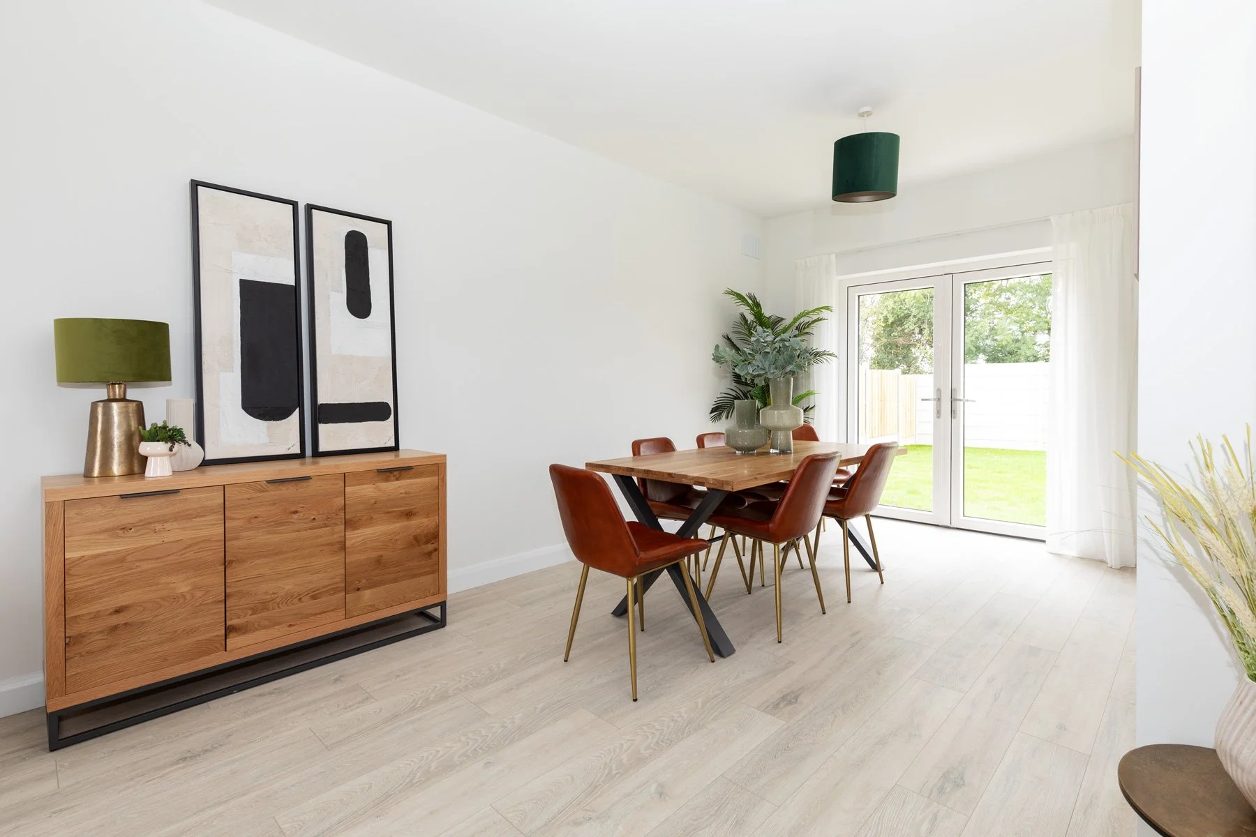 Dining room with a wooden table, six brown leather chairs, a wooden sideboard with black handles, and abstract art. Large glass doors lead to a green backyard, and there are plants and decorative items in the room.