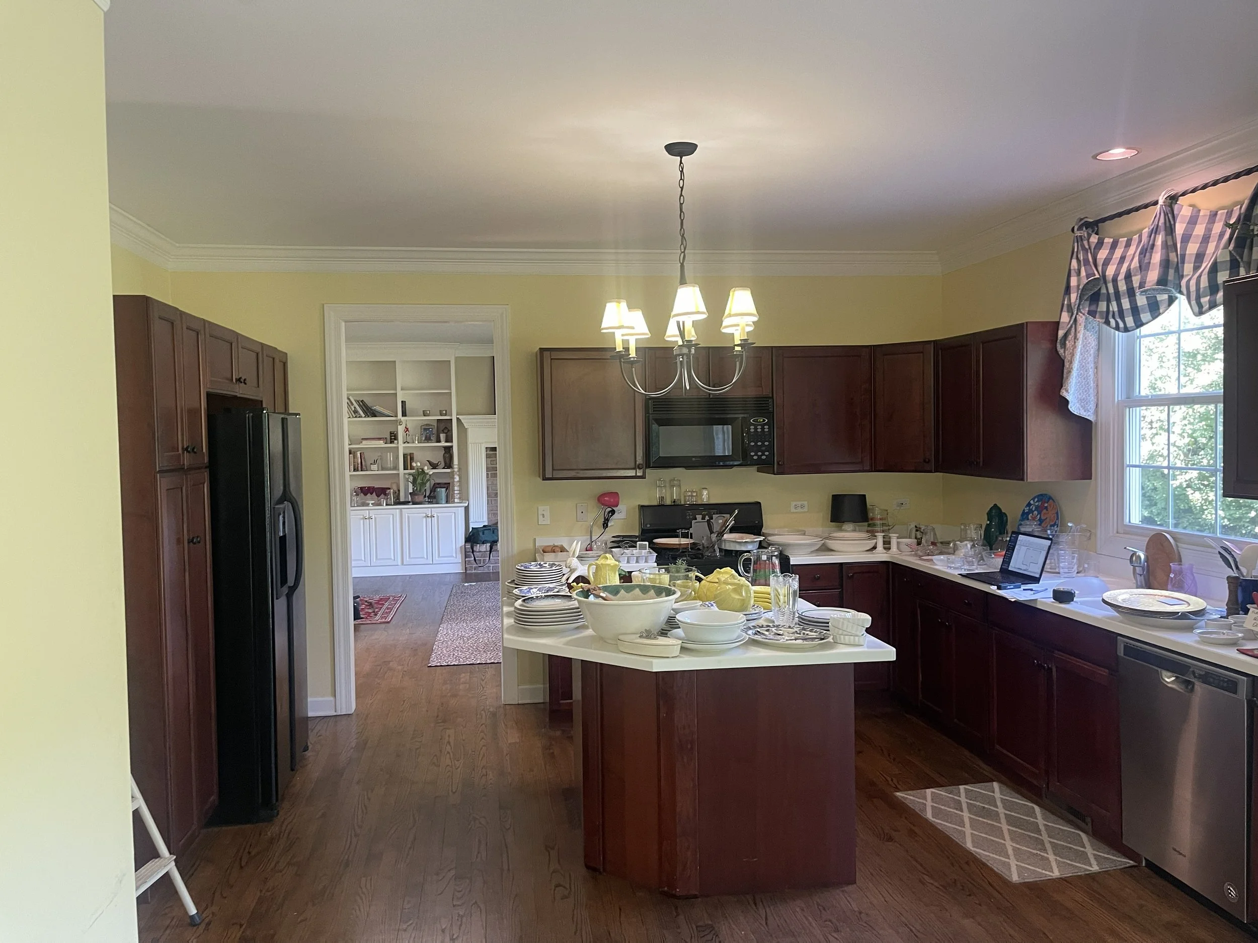 Kitchen with dark wood cabinets, a white countertop island with dishes and, a black refrigerator, and a window with a checkered valance. There are dishes and glassware on the counter and a laptop near the window.  Before pic of a kitchen remodel