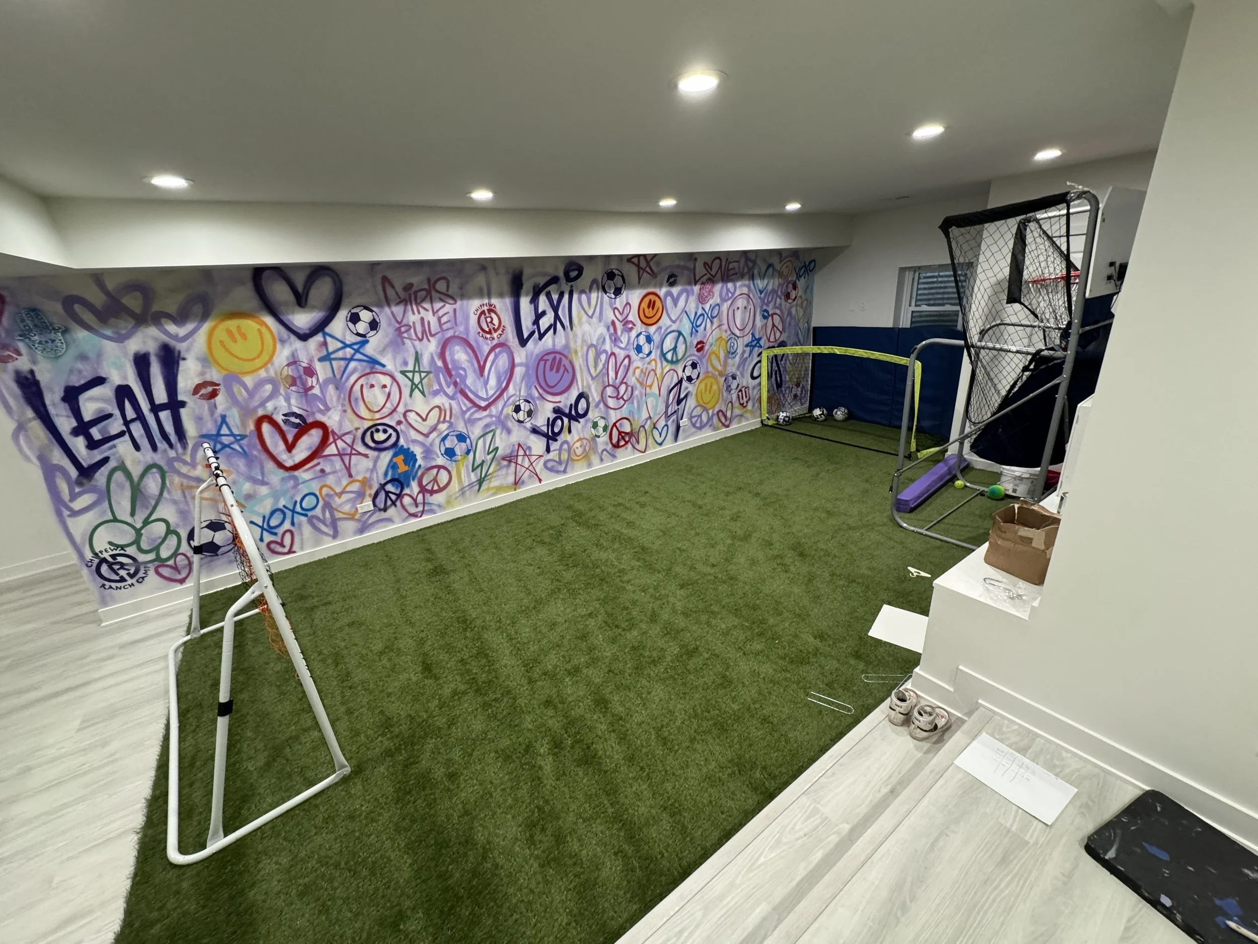 Indoor sports room as part of a basement remodel after water damage and flood with artificial turf, graffiti-covered wall, soccer goals, toys, and exercise equipment.