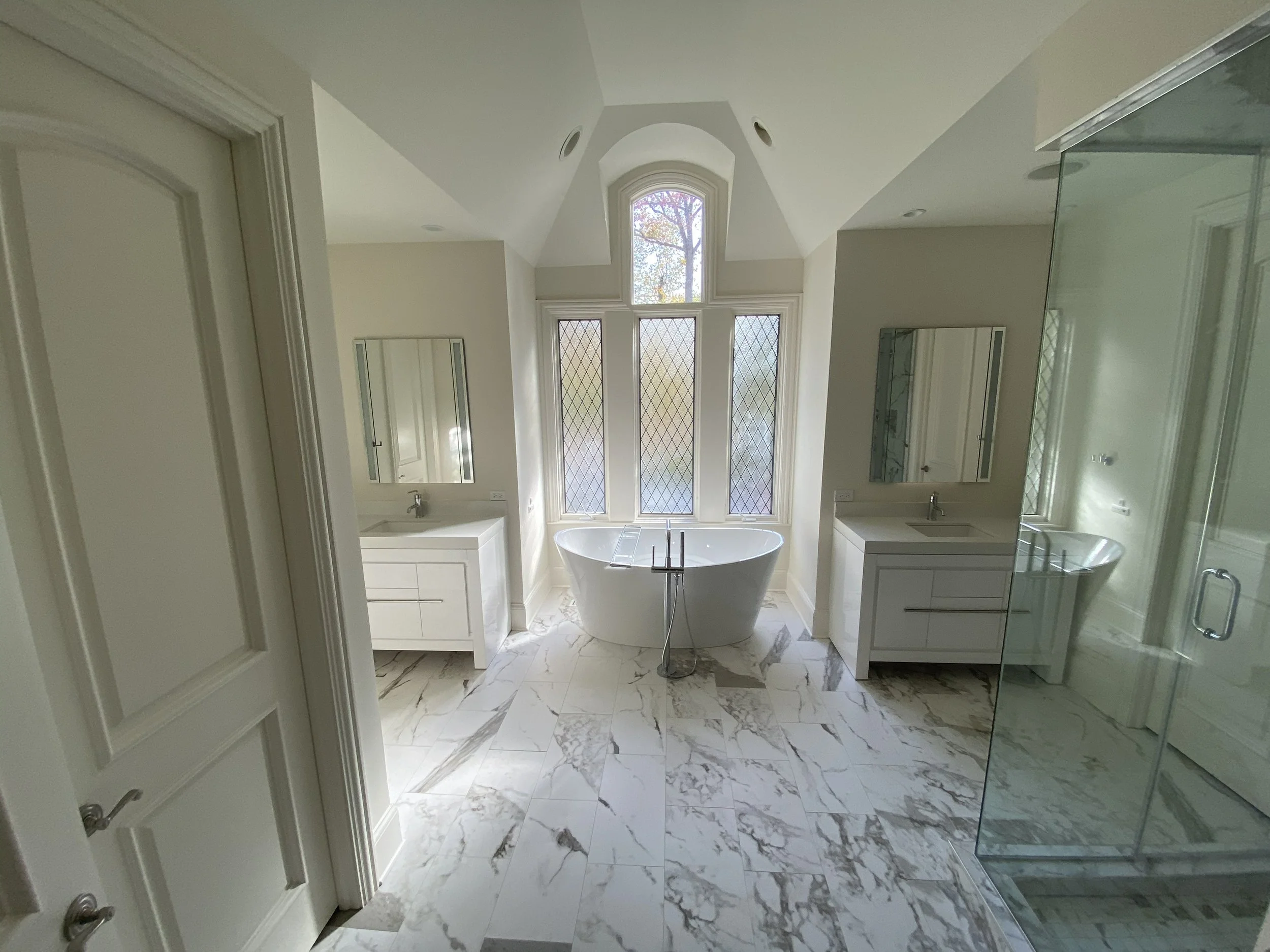 A spacious, modern bathroom remodel with a large bathtub in front of tall, decorative windows, two white vanities with mirrors, a glass shower enclosure, and marble patterned flooring.