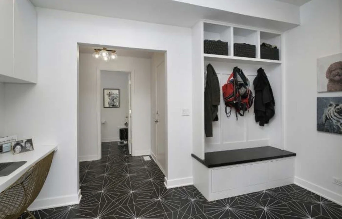 Interior view of a modern  entryway with white walls, patterned black and white geometric flooring, a built-in bench with storage, and hooks with jackets, a backpack, and bags hanging. There are shelves above the hooks and artwork on the walls.