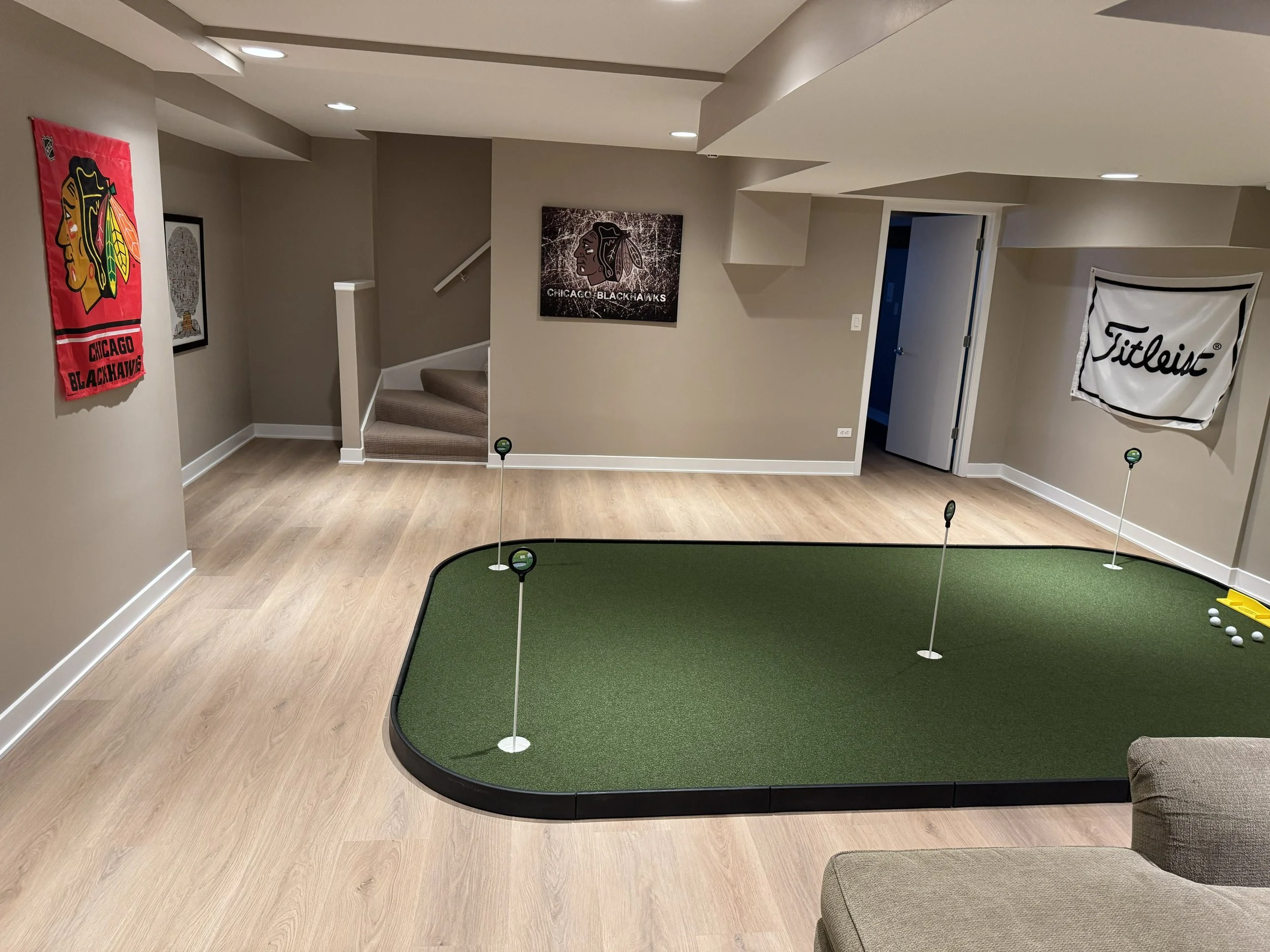 Indoor putting green with flags, golf balls, and a yellow ball holder, surrounded by wood flooring, decorative banners, and a staircase.