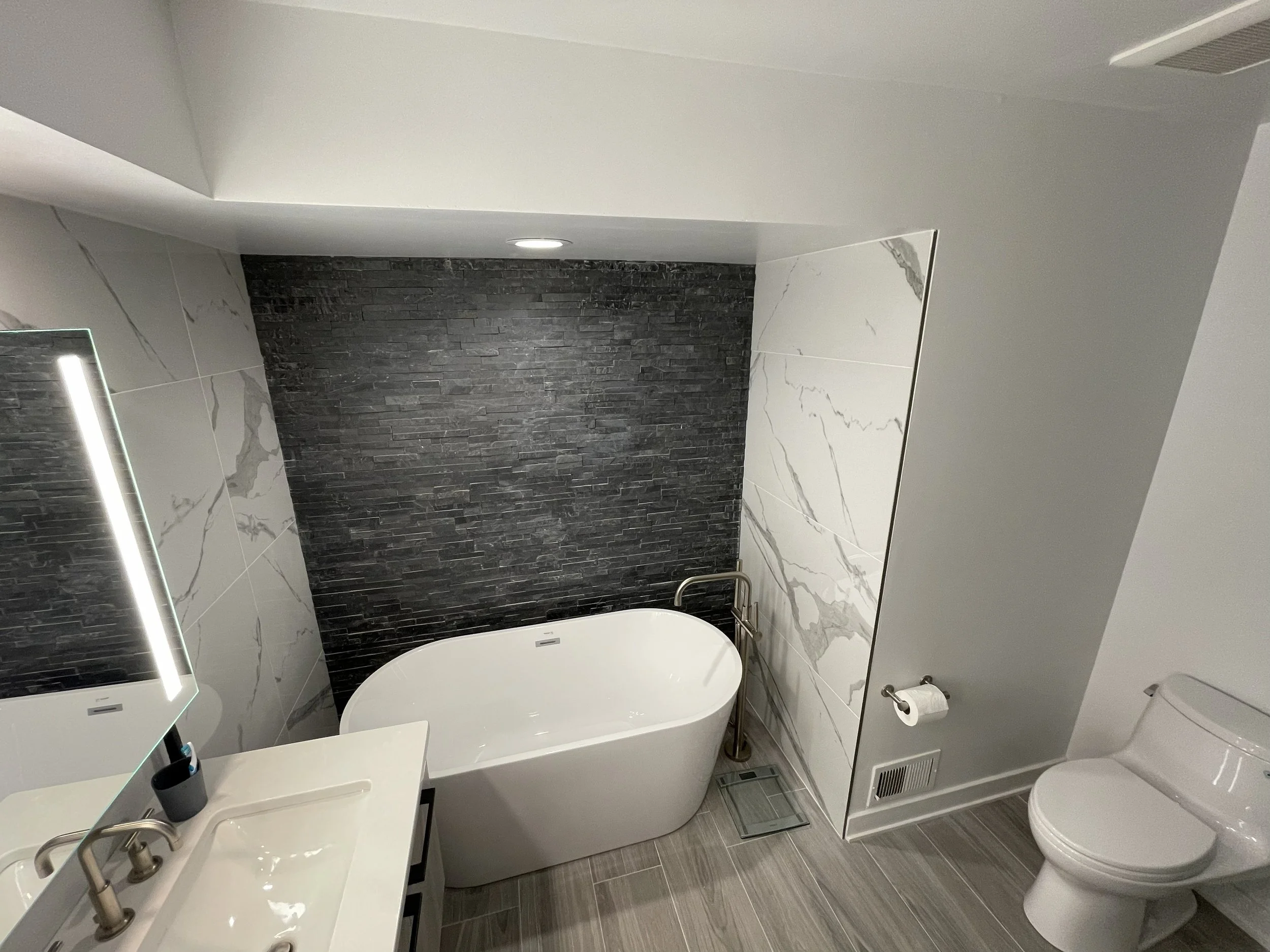 Modern bathroom remodel after a flood with a white freestanding bathtub, a vanity with a mirror, a toilet, and a dark stone accent wall.