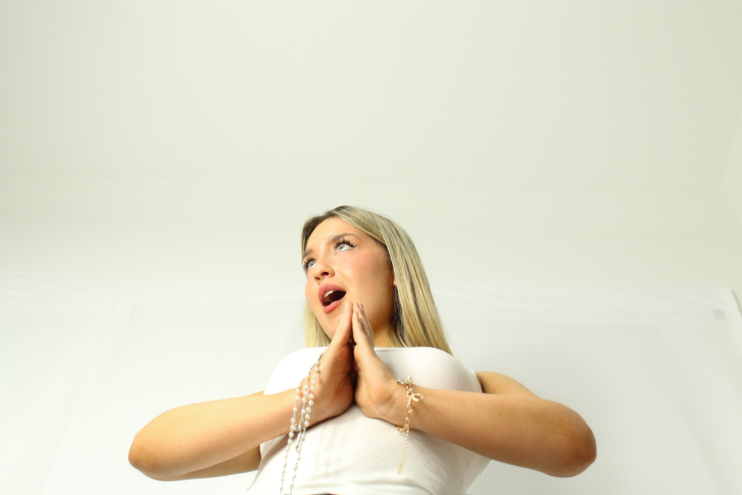 A woman with blonde hair and wearing a white top, holding a string of pearls, making a prayer-like gesture with her hands, looking upwards with a surprised or dramatic facial expression, against a plain light-colored background.