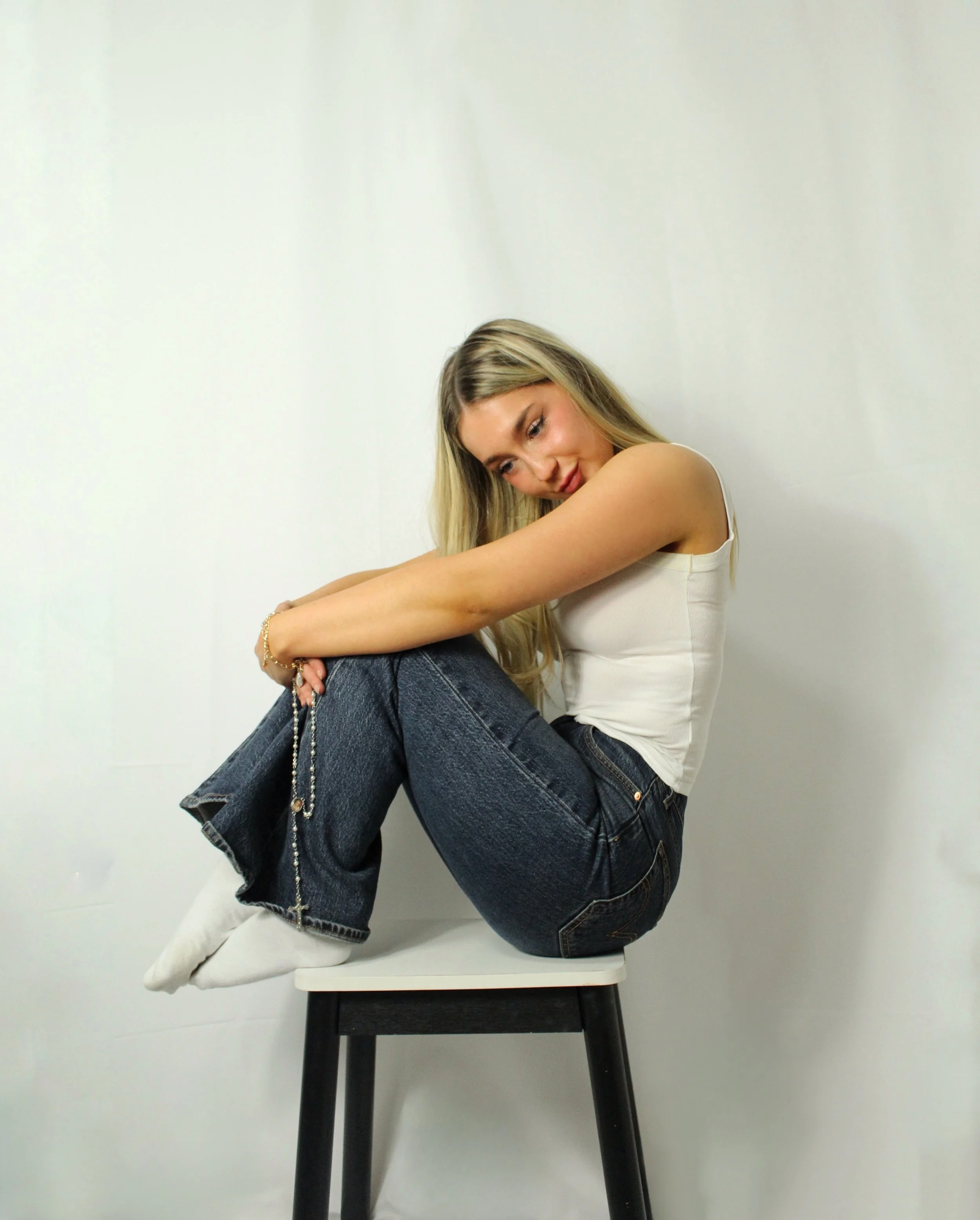 Young woman sitting on a stool with her legs crossed, wearing a white tank top, jeans, and white socks, against a plain white background.
