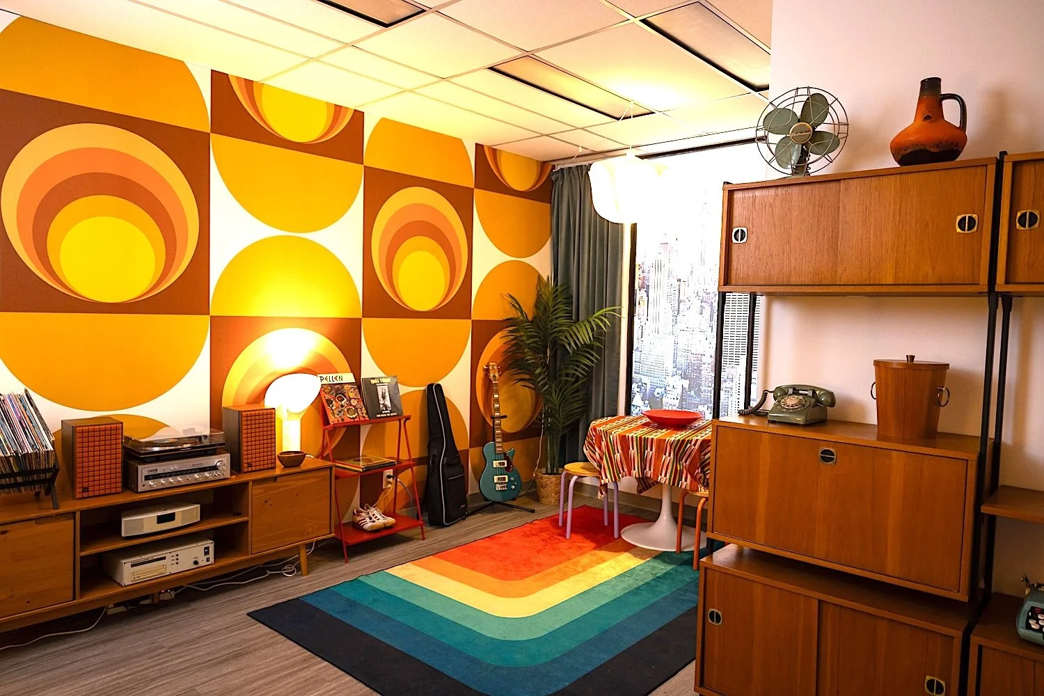 Living room with colorful 1970s-style decor, featuring a rainbow-striped rug, wooden furniture, an electric guitar, vinyl records, and a large window with city view.